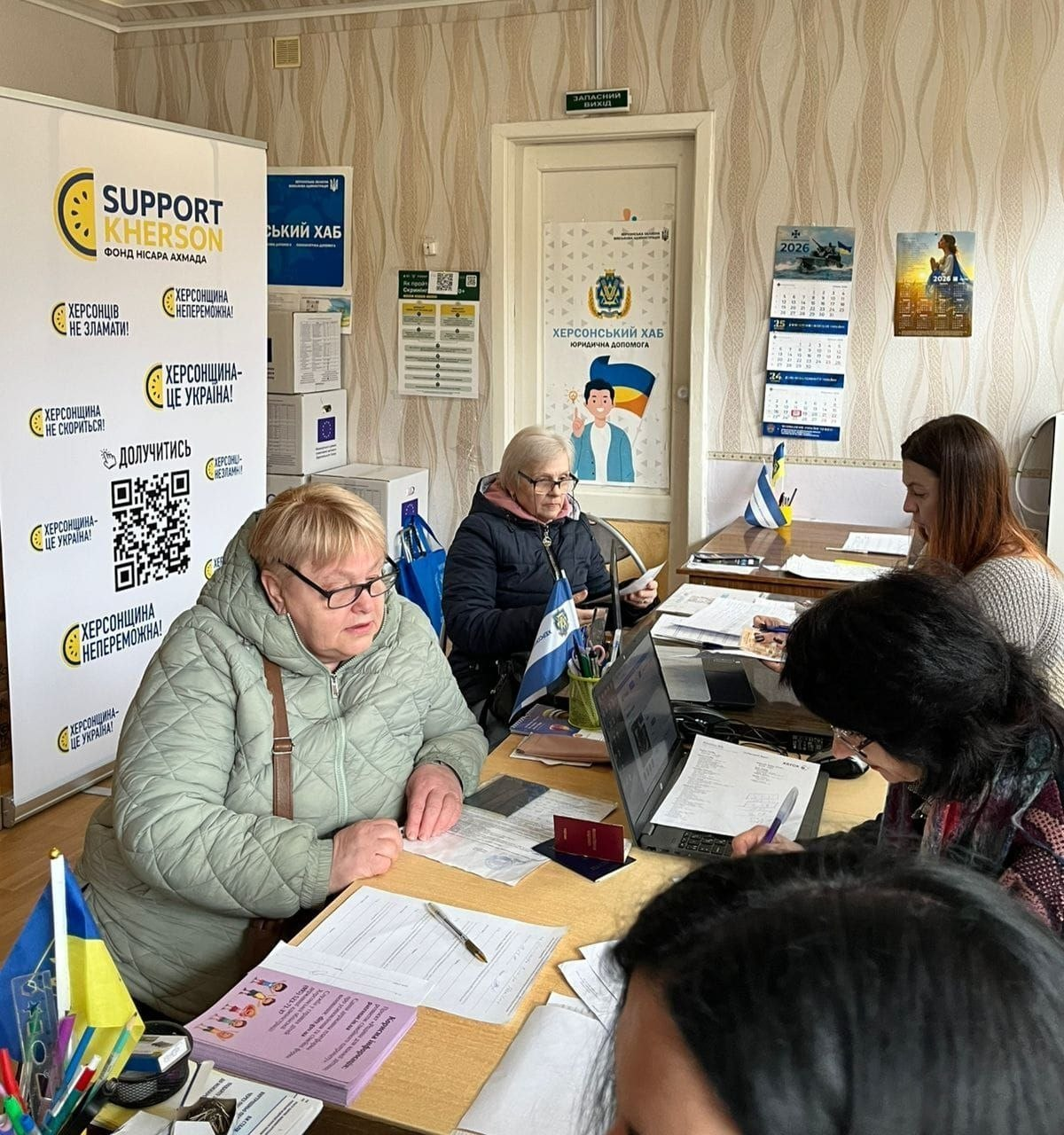 Жінки в офісі «Херсонського хабу» працюють із документами за столами з ноутбуком та прапорцями. Поруч стоїть великий банер «Support Kherson» із патріотичними гаслами «Херсонщина — це Україна!» та «Херсонців не зламати!».