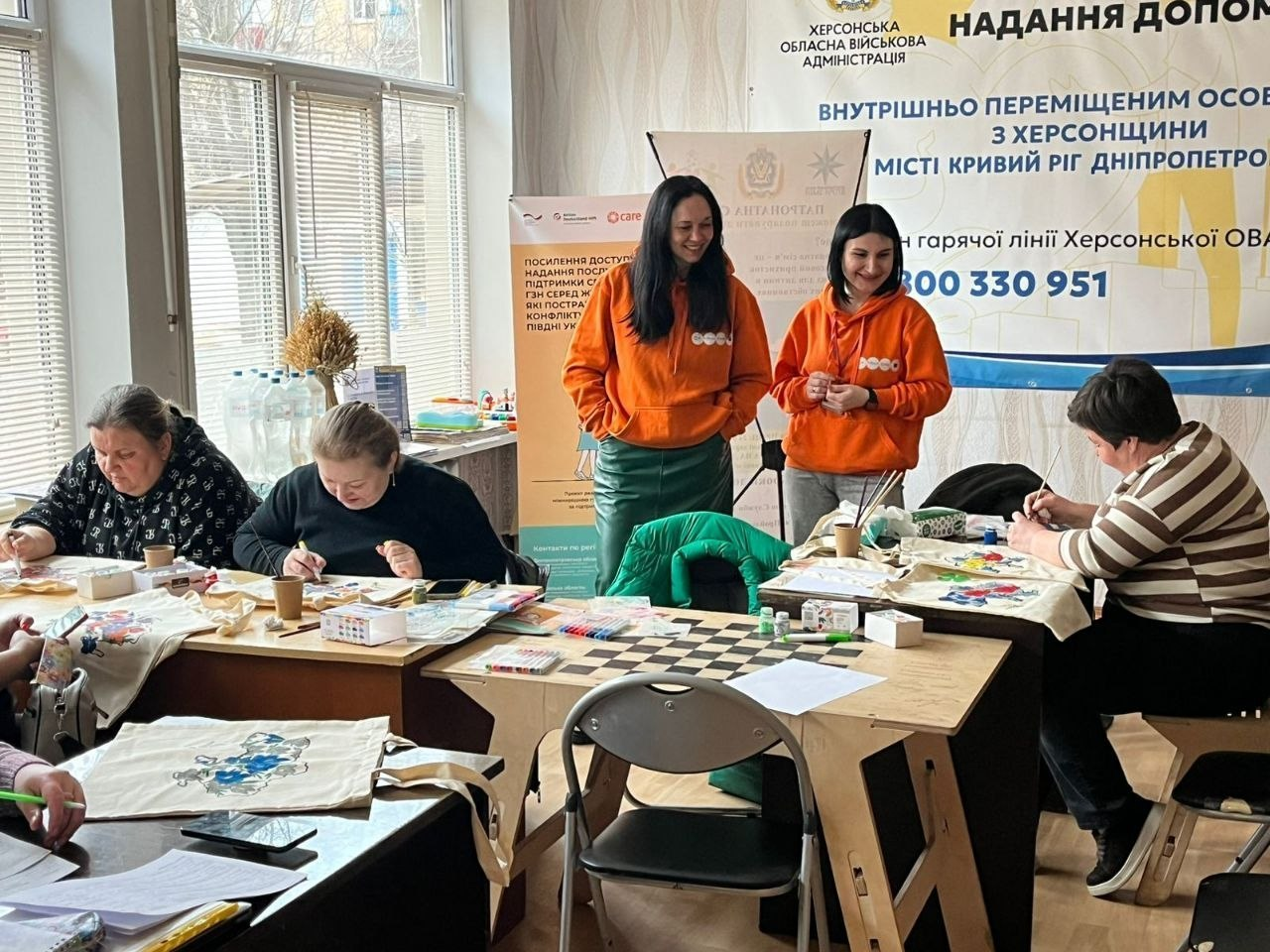 У світлому приміщенні жінки розписують екосумки фарбами, сидячи за столами. Поруч стоять дві дівчини в яскраво-помаранчевих худі та посміхаються. На фоні видно великий інформаційний банер Херсонської ОВА з контактами гарячої лінії для допомоги ВПО у Кривому Розі.