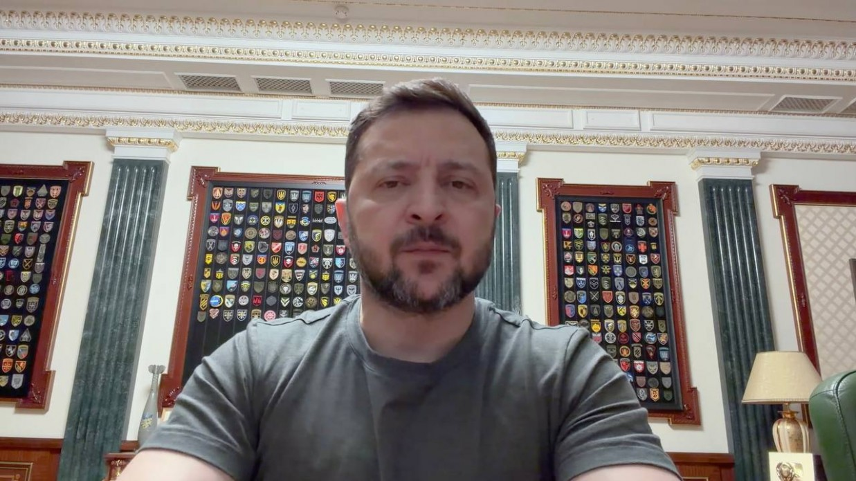 Чоловік у футболці кольору хакі на фоні кабінету зі стендами, заповненими численними військовими шевронами та емблемами.