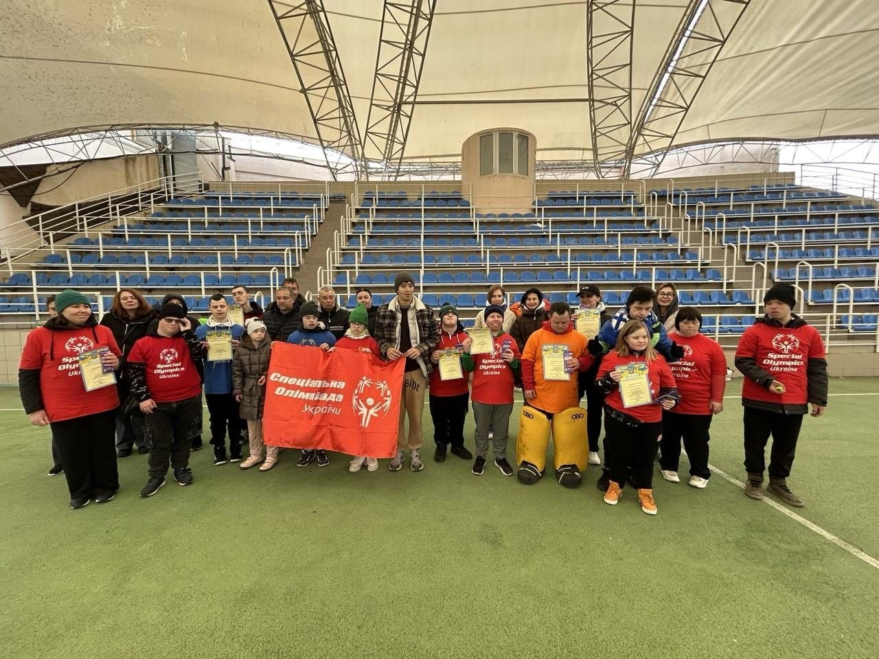 Велика група учасників «Спеціальної Олімпіади України» позує для спільного фото на спортивному майданчику. Багато хто з них у червоних фірмових футболках, дехто тримає дипломи та медалі, а попереду розгорнуто великий червоний прапор організації на фоні синіх трибун стадіону.