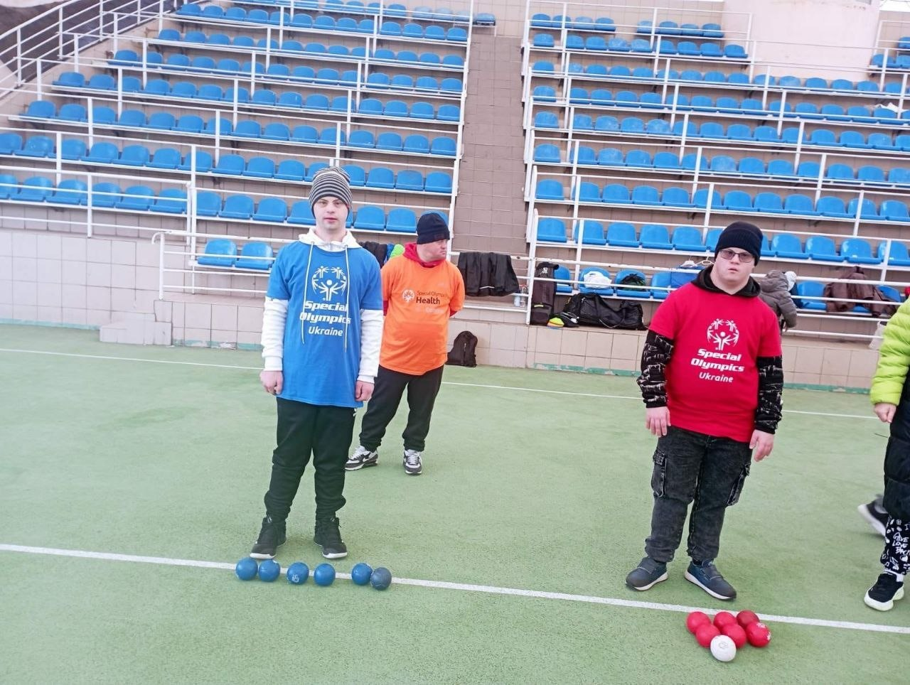 Троє спортсменів у фірмових футболках «Спеціальної Олімпіади України» грають у бочче на відкритому майданчику на фоні порожніх синіх трибун стадіону. Перед ними на полі лежать сині та червоні кулі.