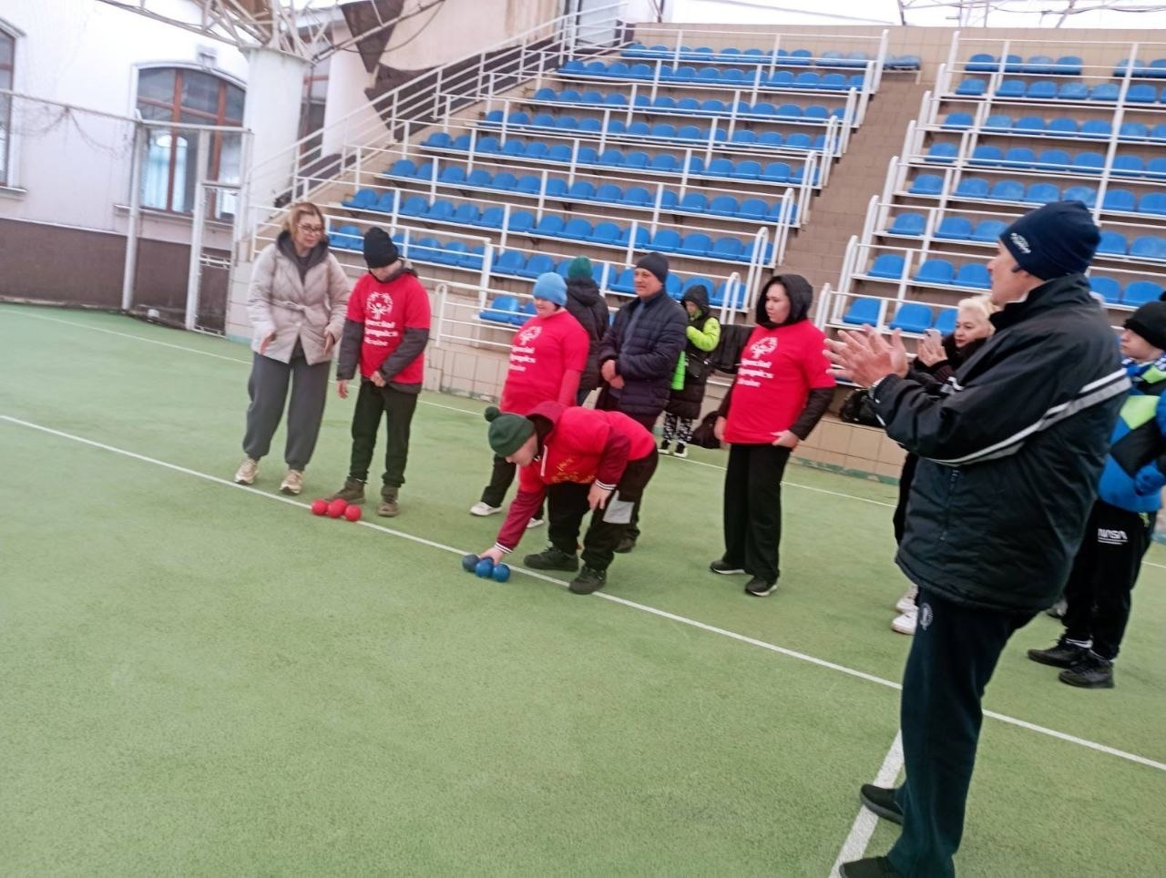 Група людей на спортивному майданчику грає в бочче на фоні синіх трибун. Учасник у червоній футболці готується кинути кулю, а інші спостерігають за грою та аплодують.