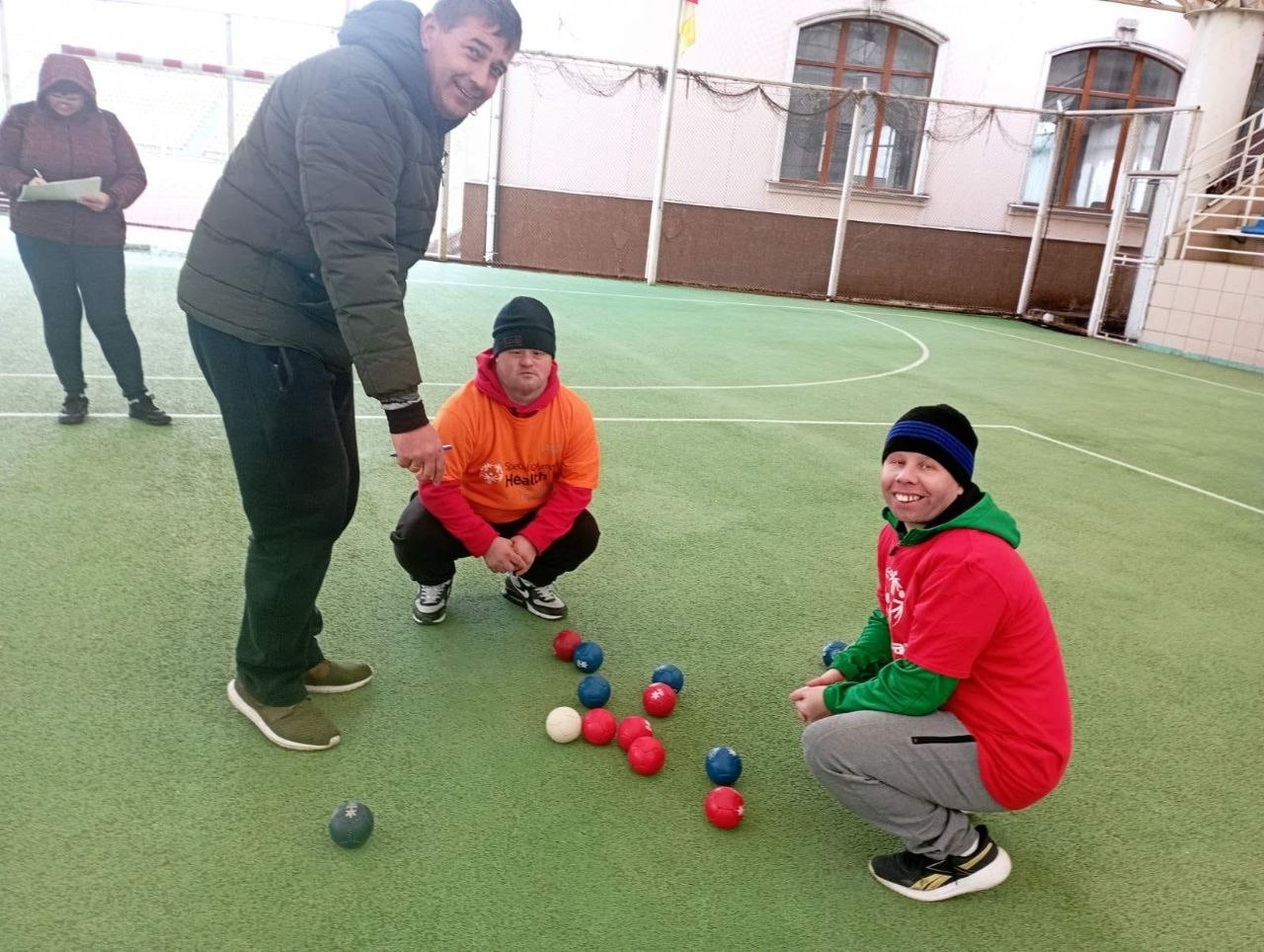 Троє чоловіків на зеленому спортивному майданчику грають у бочче. Двоє гравців у яскравих худі присіли біля червоних, синіх та білої куль, а чоловік поруч із ними стоїть і спостерігає за грою.