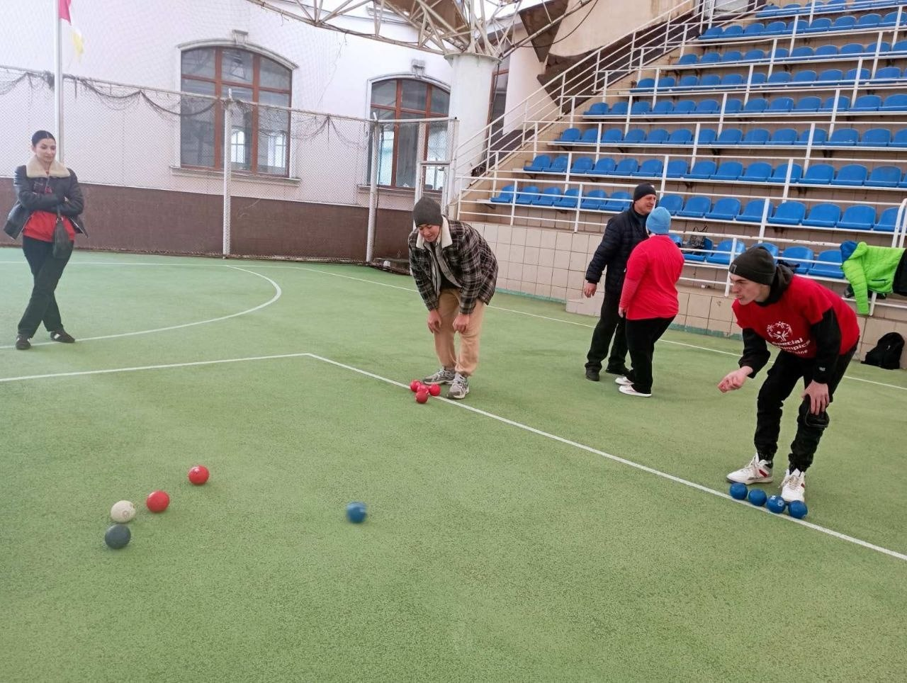 Група людей на спортивному майданчику грає в бочче. Один з учасників у червоній футболці готується до кидка, а на зеленому покритті поля лежать розкидані сині, червоні та білі кулі.