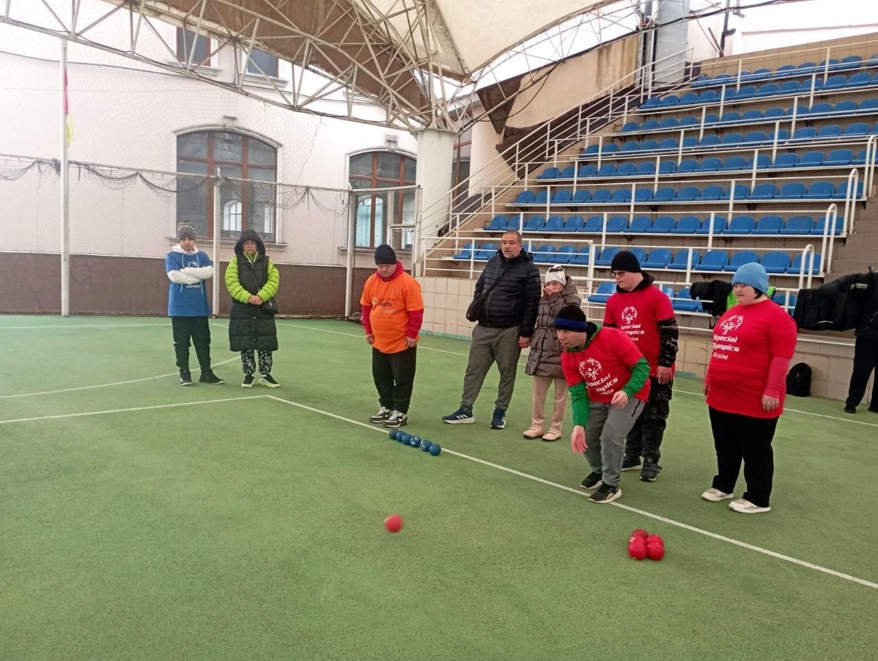 Група людей на спортивному майданчику грає в бочче на фоні синіх трибун. Учасник у червоній футболці з емблемою «Спеціальної Олімпіади» нахилився для кидка червоної кулі, а інші спостерігають за процесом.