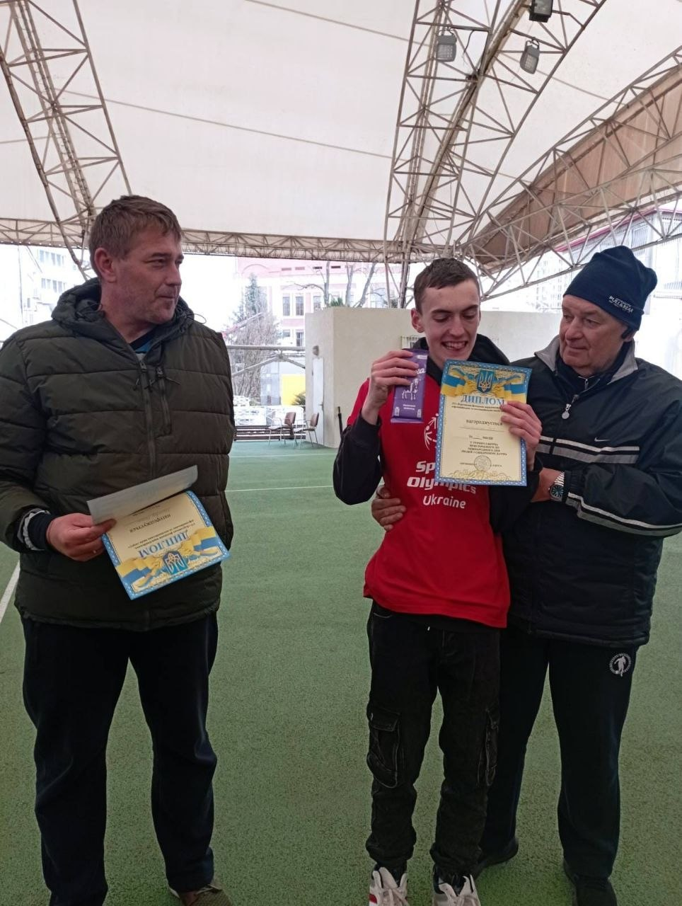 Троє чоловіків на критому спортивному майданчику під час нагородження. Учасник у червоній футболці з написом Special Olympics Ukraine радісно тримає диплом і медаль, а двоє чоловіків поруч підтримують його, також тримаючи дипломи.