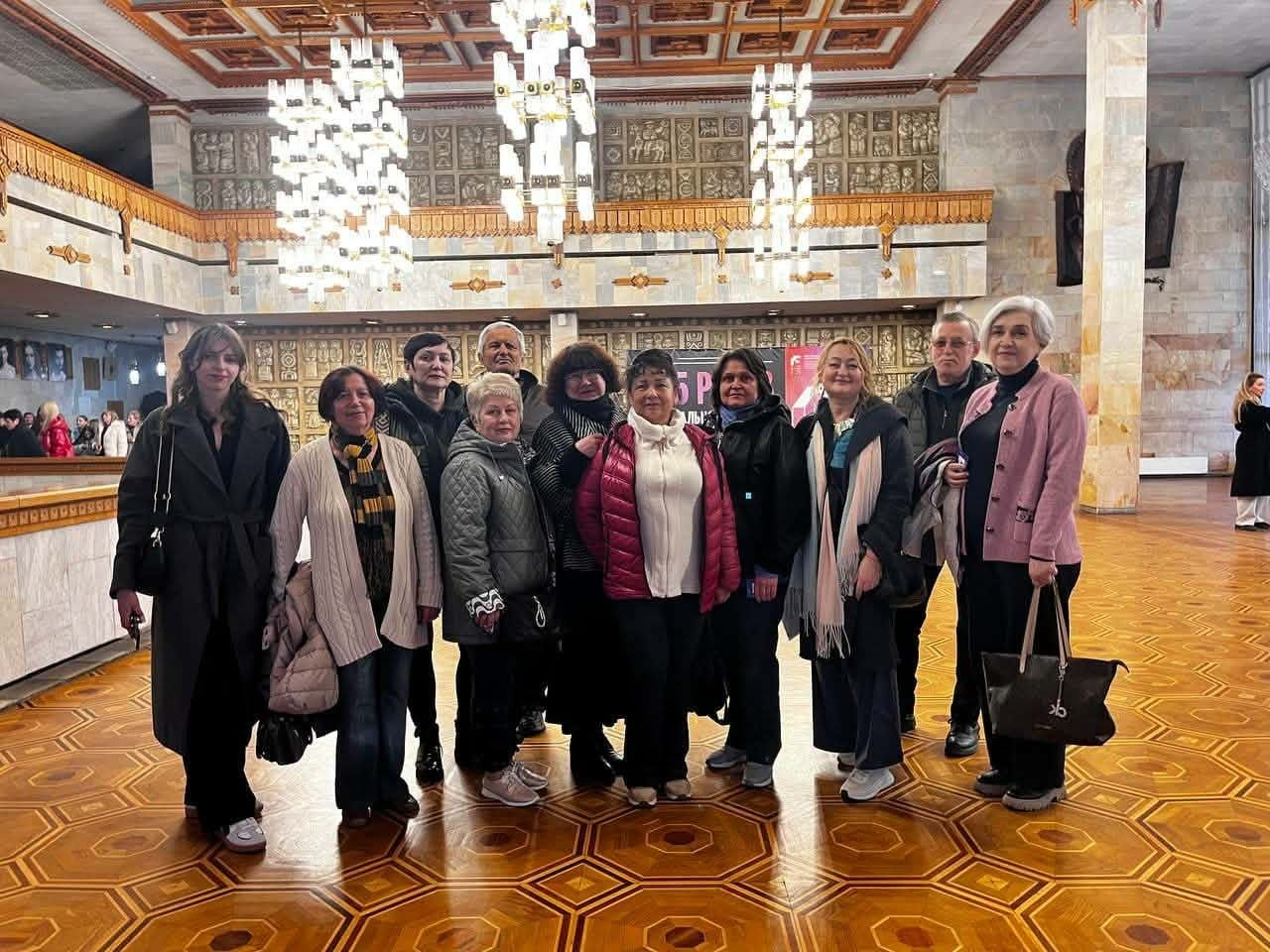 Група людей у верхньому одязі позує для фото у просторому фойє театру з візерунчастим паркетом. На фоні видно високі колони, декоративні стіни та масивні кришталеві люстри.