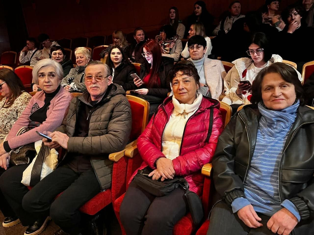Група глядачів різного віку в теплих куртках сидить у театральному залі з червоними кріслами. На передньому плані четверо осіб дивляться в камеру, за ними видно інші ряди людей у затіненому приміщенні.