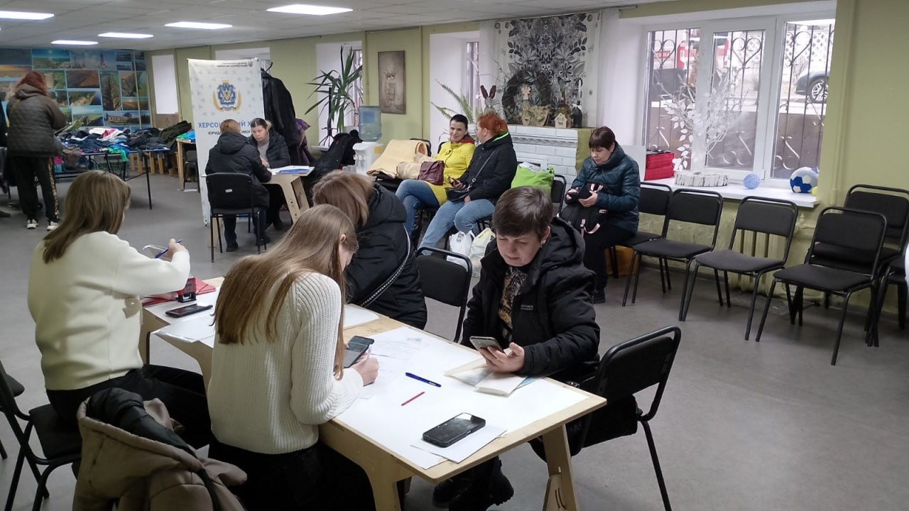 Група людей у світлому приміщенні гуманітарного або інформаційного центру. Кілька осіб сидять за столами та заповнюють документи, інші очікують своєї черги на стільцях уздовж стіни. На фоні видно банер з гербом України та стоси речей.