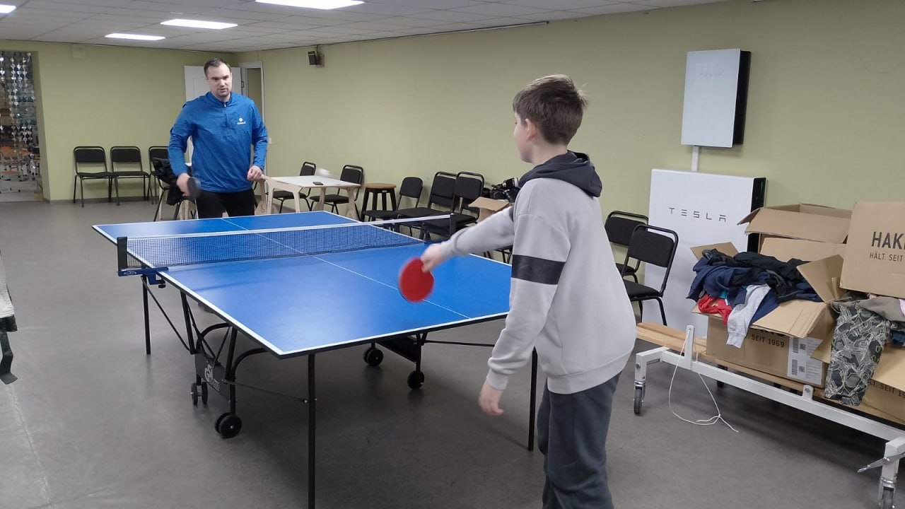 Чоловік та підліток грають у настільний теніс за синім столом у просторому приміщенні. Поруч стоять коробки з речами та обладнання Tesla, а на фоні видно ряди стільців уздовж стіни.