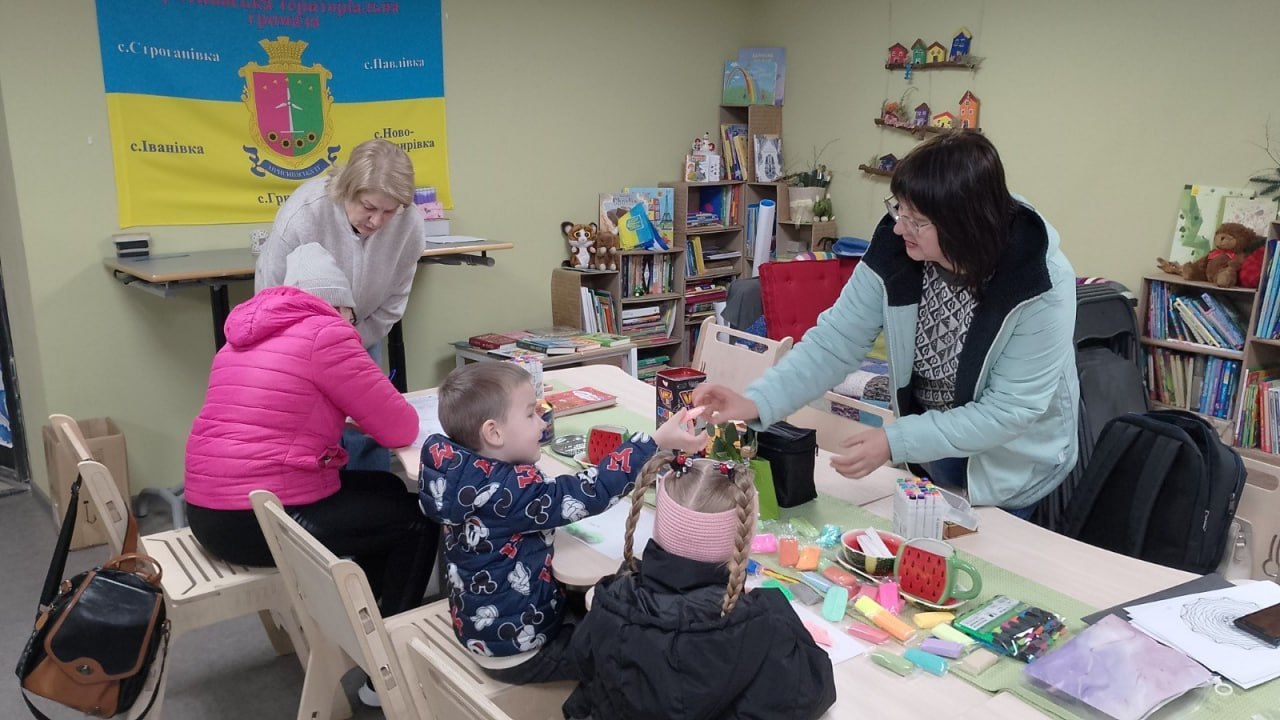 Двоє дітей та троє дорослих займаються творчістю за столом у дитячому куточку або бібліотеці. На столі лежать фломастери та розмальовки, а на фоні — полиці з книжками та жовто-блакитний прапор із гербом громади.