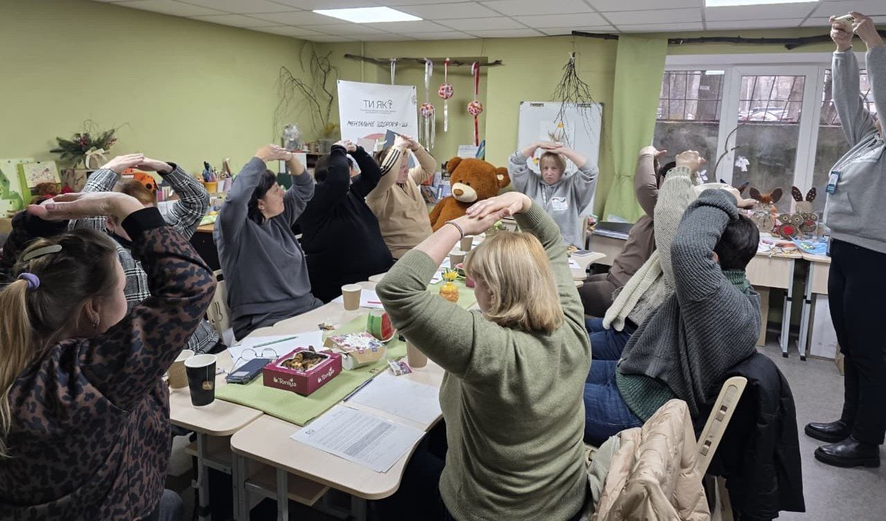 Група жінок сидить за столом у світлій кімнаті та виконує психологічну вправу, склавши руки над головою у формі даху. На фоні видно плакат із написом «Ти як?», великого іграшкового ведмедя та канцелярське приладдя на столах.