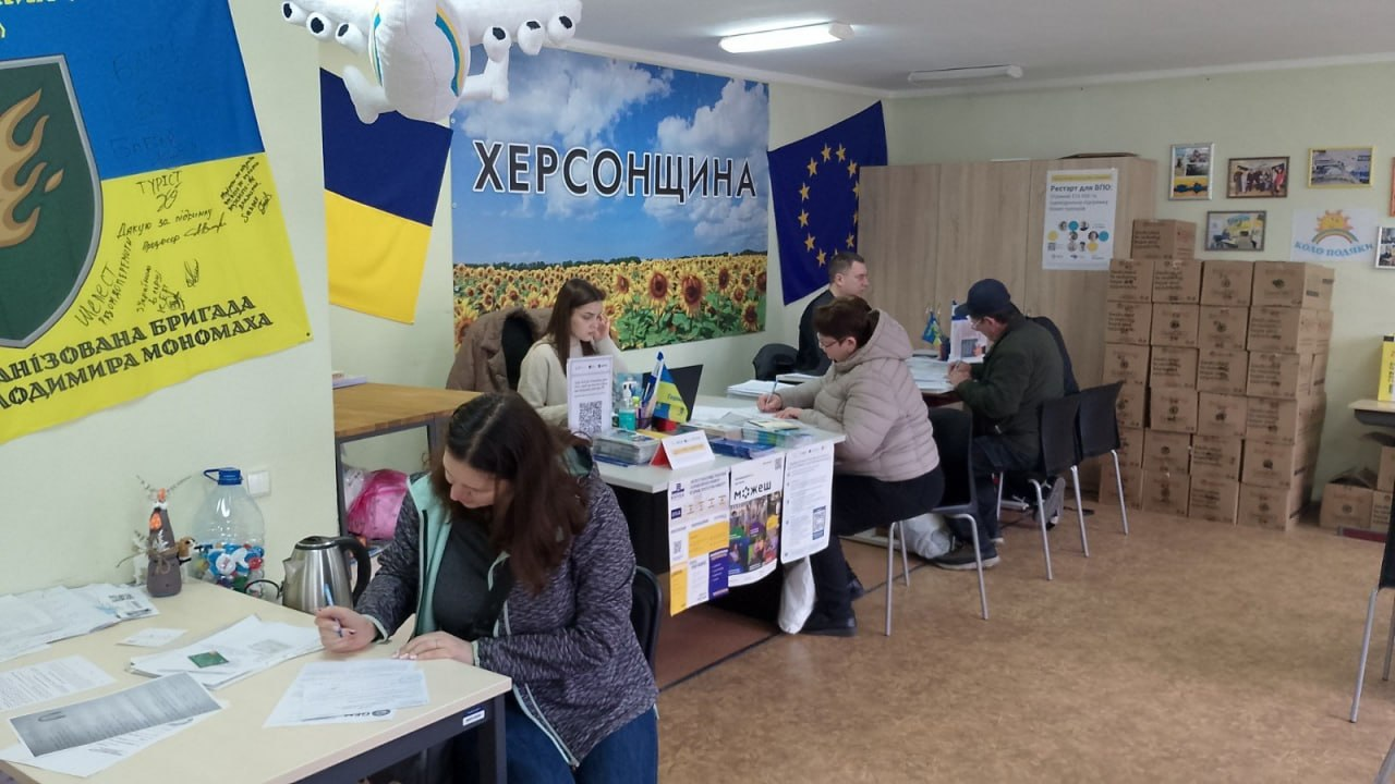 Робоче приміщення гуманітарного штабу з прапорами України та ЄС на стінах. Люди за столами заповнюють документи, а поруч складено стоси картонних коробок.