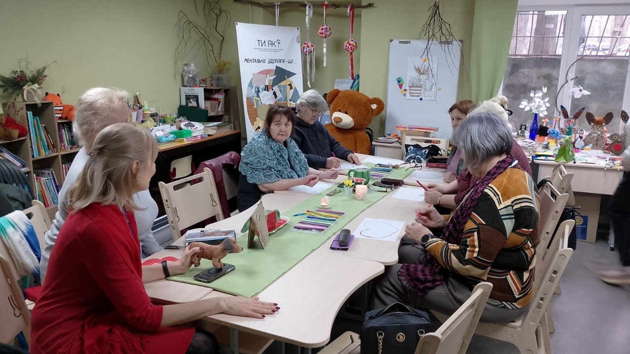 Група жінок сидить за великим столом у затишній кімнаті під час заняття з арттерапії. На столі лежать олівці та малюнки, а на фоні видно великого плюшевого ведмедя та банер програми ментального здоров'я «Ти як?».
