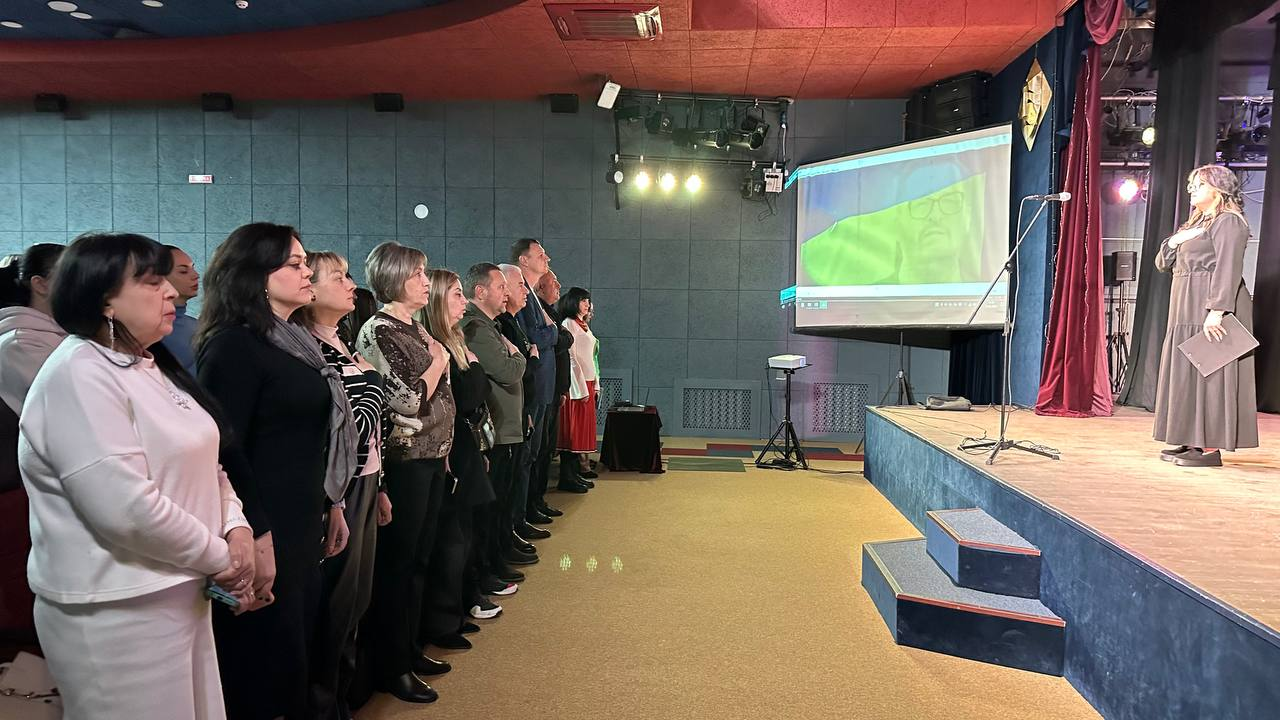 Група людей стоїть у залі, приклавши праву руку до серця, під час урочистої події. Вони повернуті до сцени, де на великому екрані транслюється відео, а поруч стоїть жінка-ведуча. Атмосфера виглядає офіційною та шанобливою.