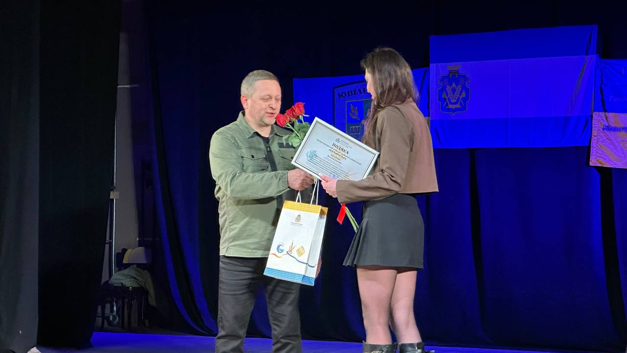 На сцені чоловік у військовому одязі вручає молодій жінці грамоту, букет червоних троянд та подарунковий пакет. Позаду них на синьому фоні розміщено прапори та офіційну символіку.