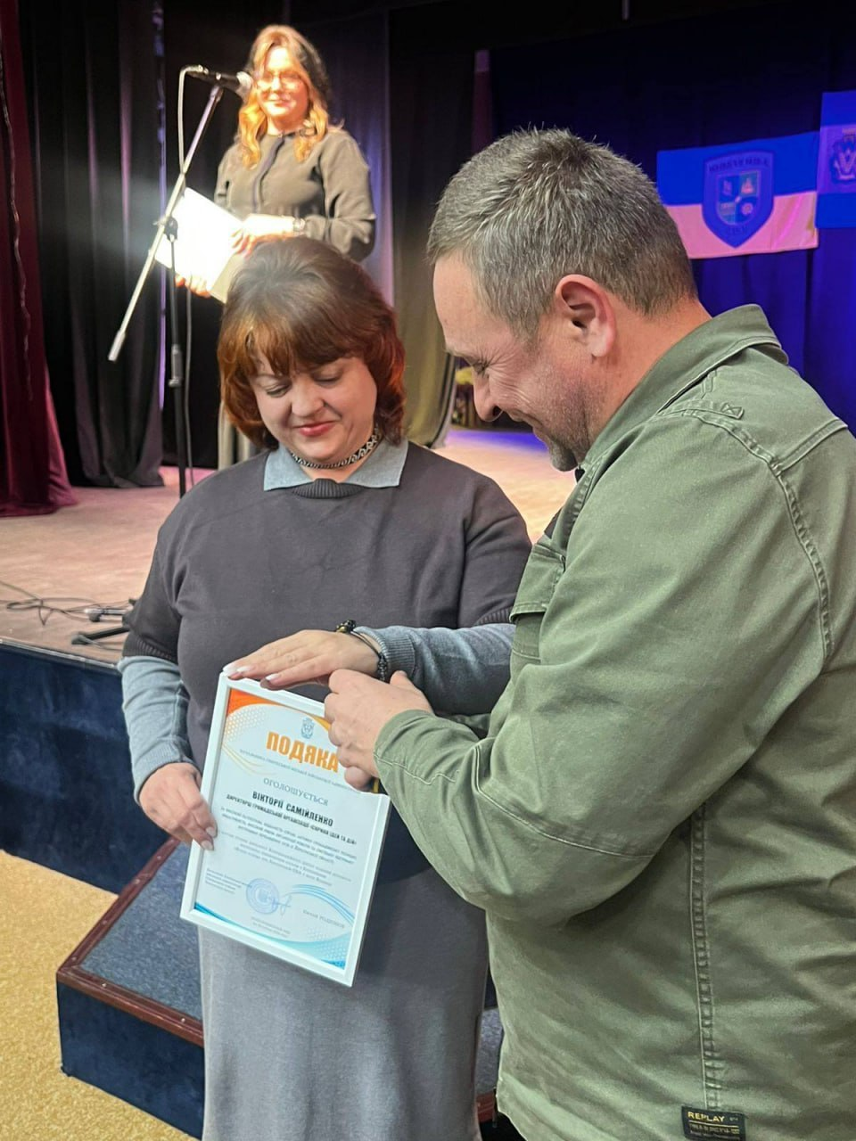 Чоловік вручає жінці офіційну подяку в рамці під час урочистої події. На задньому плані видно сцену з ведучою та національну символіку. Обидва учасники посміхаються, створюючи святкову атмосферу.