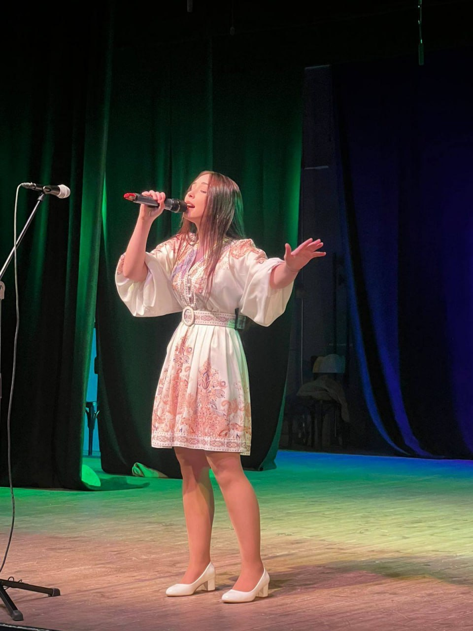 Дівчина співає на сцені в мікрофон під час виступу. Вона одягнена у світлу сукню з українським орнаментом, широкими рукавами та поясом. Сцена освітлена кольоровими прожекторами на фоні темно-зелених завіс.