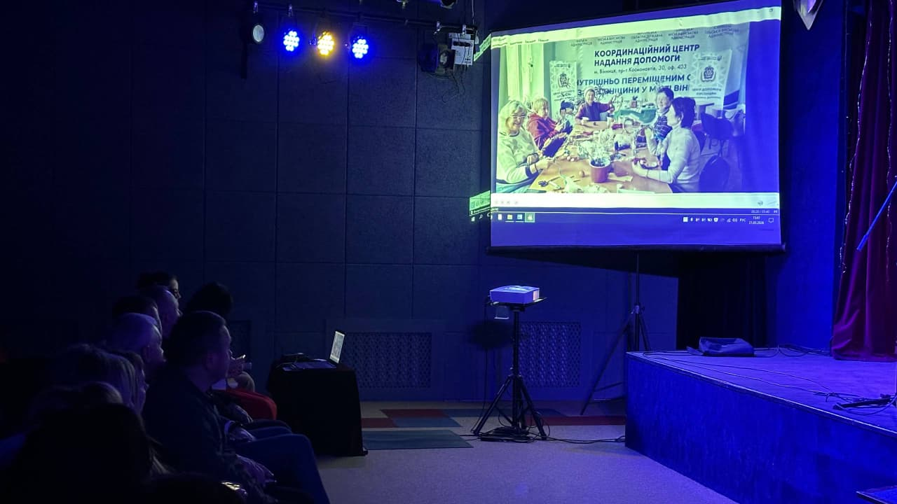 Глядачі в темній залі дивляться на великий екран, де за допомогою проектора транслюється презентація про Координаційний центр надання допомоги. На екрані видно фотографію групи людей за столом. Сцена підсвічена синіми та жовтими прожекторами.