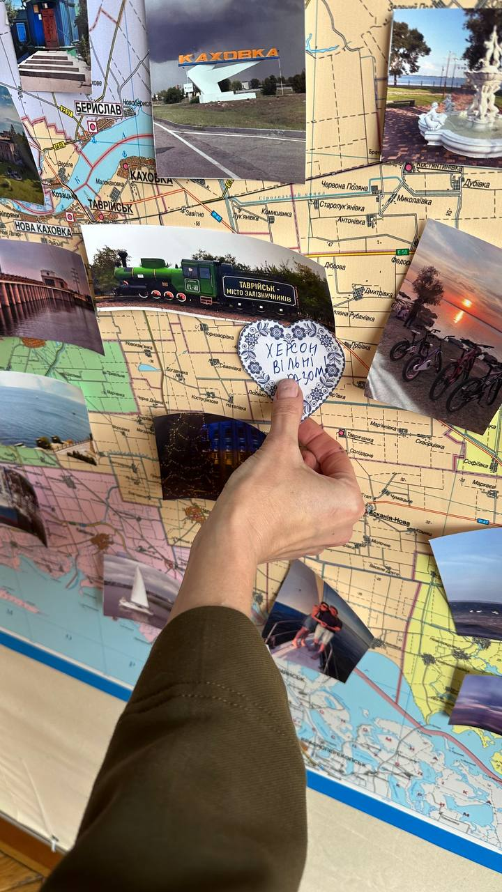 Рука людини кріпить паперове серце з написом «Херсон вільні разом» на велику карту Херсонської області. Навколо розміщено фотографії визначних місць регіону: Каховка, Берислав, Таврійськ та інші місцеві краєвиди.