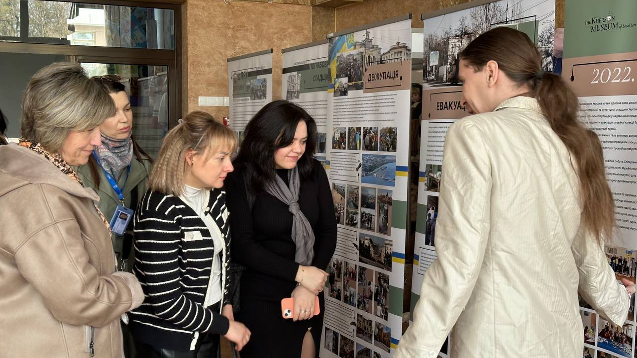 Група жінок уважно розглядає інформаційні стенди мобільної виставки. Стенди містять фотографії та тексти про події 2022 року, зокрема з розділами «Деокупація» та «Евакуація». Жінка праворуч, повернута спиною до камери, імовірно, проводить екскурсію або надає пояснення.