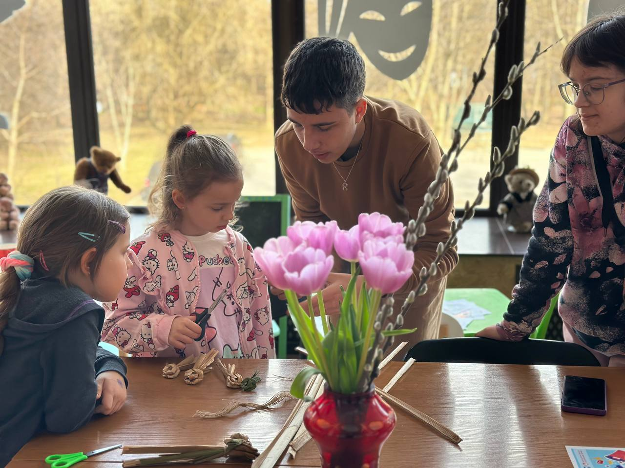 Група дітей та молода людина займаються творчістю за столом у світлому приміщенні. Вони виготовляють вироби з природних матеріалів (соломи або лози). На передньому плані стоїть ваза з рожевими тюльпанами та гілками верби, що створює весняну атмосферу.