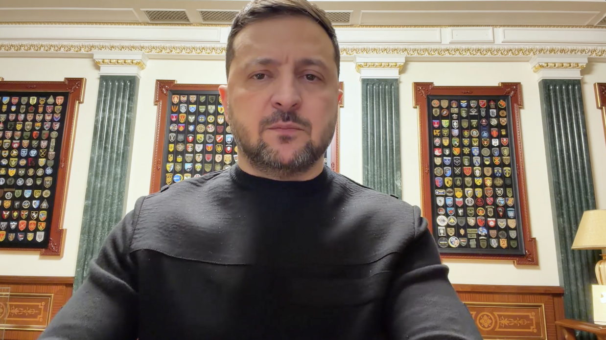 Чоловік у чорному светрі звертається до аудиторії під час відеозвернення. Він знаходиться в офіційному кабінеті, на фоні якого видно колони та стенди з численними військовими шевронами та емблемами.