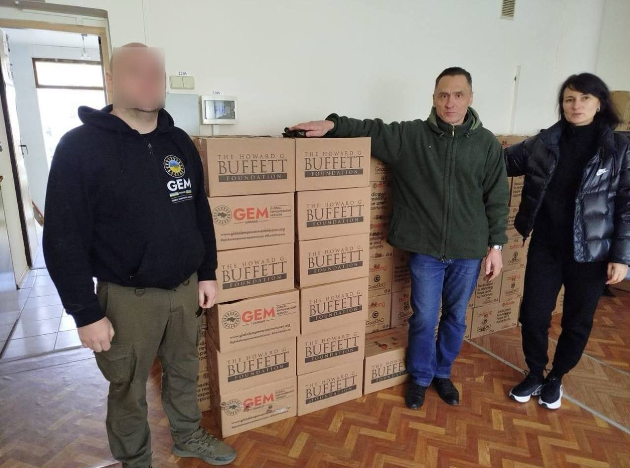 Три особи стоять біля великої кількості коробок з гуманітарною допомогою від фондів GEM та The Howard G. Buffett Foundation у приміщенні з паркетною підлогою.