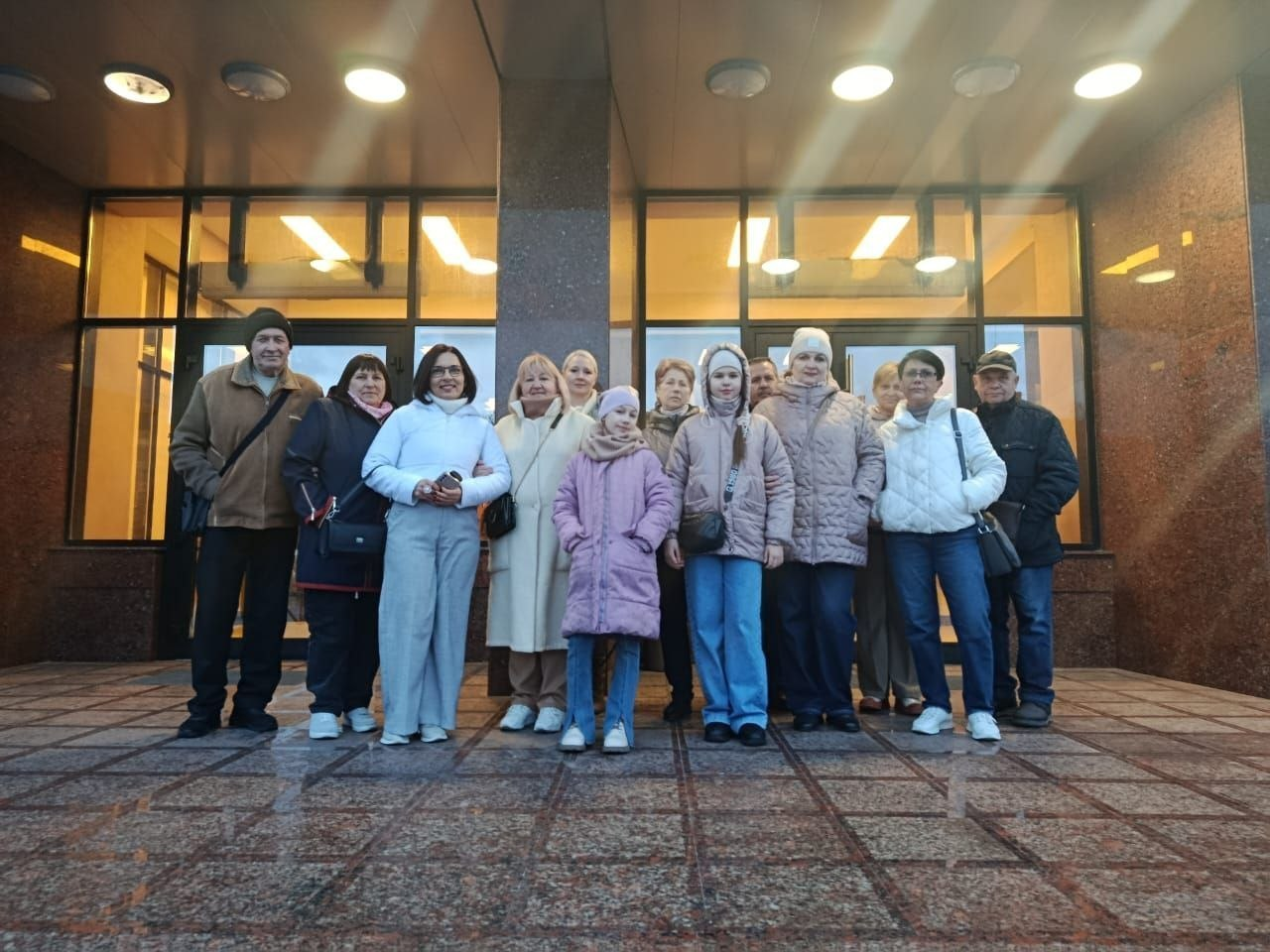 Група людей різного віку в теплому одязі позує для спільного фото перед входом до сучасної будівлі з великими скляними дверима та яскравим освітленням.