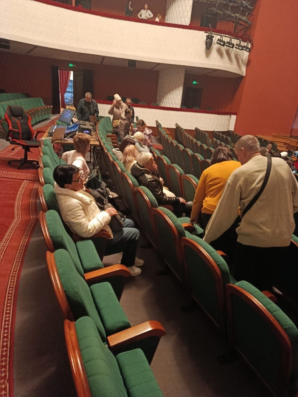 Глядацька зала театру або кінотеатру із зеленими кріслами, в якій розмістилася невелика група людей у верхньому одязі. Зліва видно робоче місце звукорежисера з обладнанням.