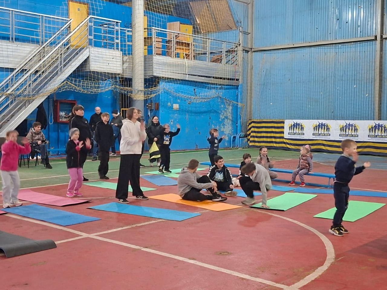 Група дітей та підлітків проводить тренування у спортивному залі з синіми стінами. Частина дітей займається на кольорових килимках, інші стоять поруч; на фоні видно банер «Спорт для всіх».