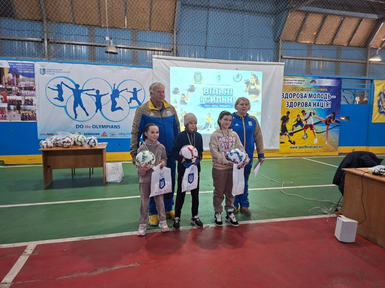 Троє дітей та двоє дорослих у спортивній формі позують у спортзалі під час нагородження. Діти тримають у руках футбольні м’ячі та подарункові пакети на фоні тематичних банерів про спорт.