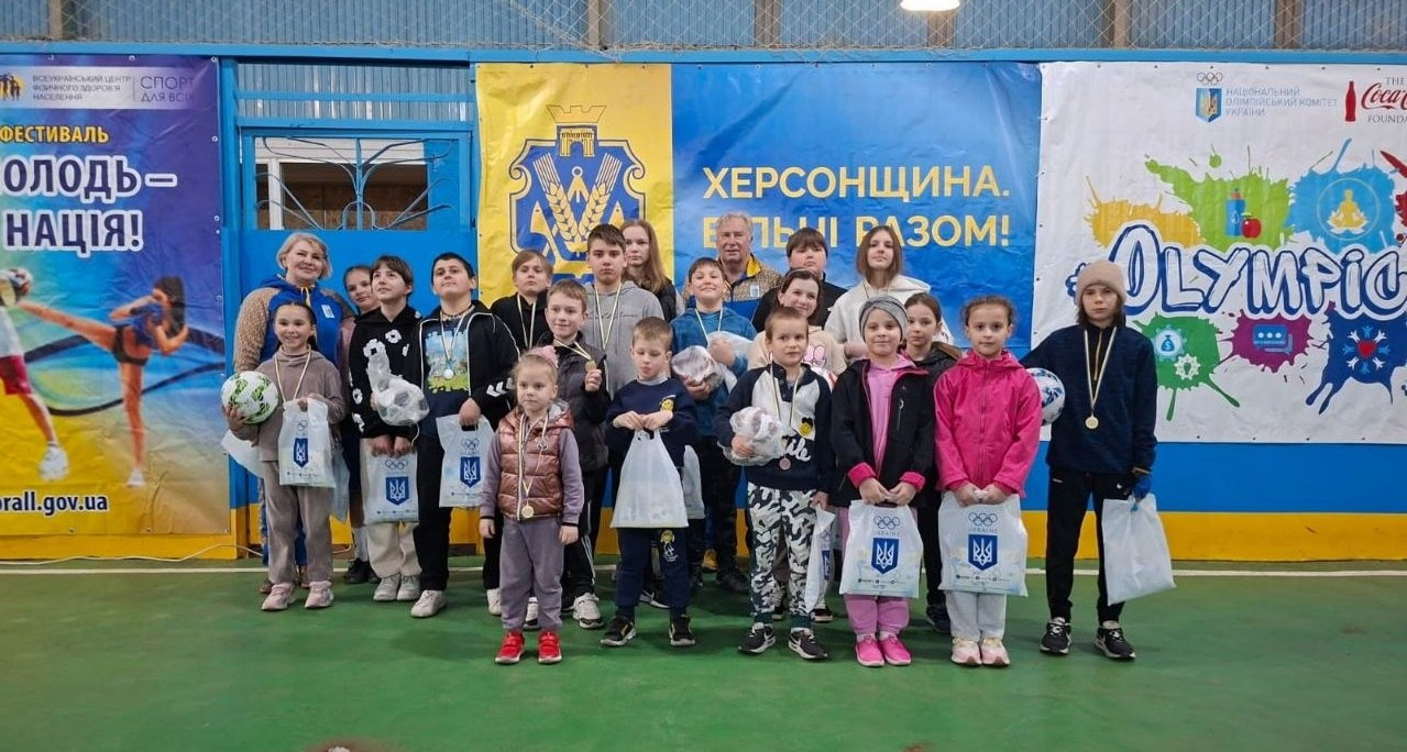 Велика група дітей та дорослих у спортивному залі позує для спільного фото після заходу. Діти тримають подарунки та м'ячі; на фоні розміщено патріотичні банери з написом «Херсонщина. Вільні разом!» та олімпійською символікою.