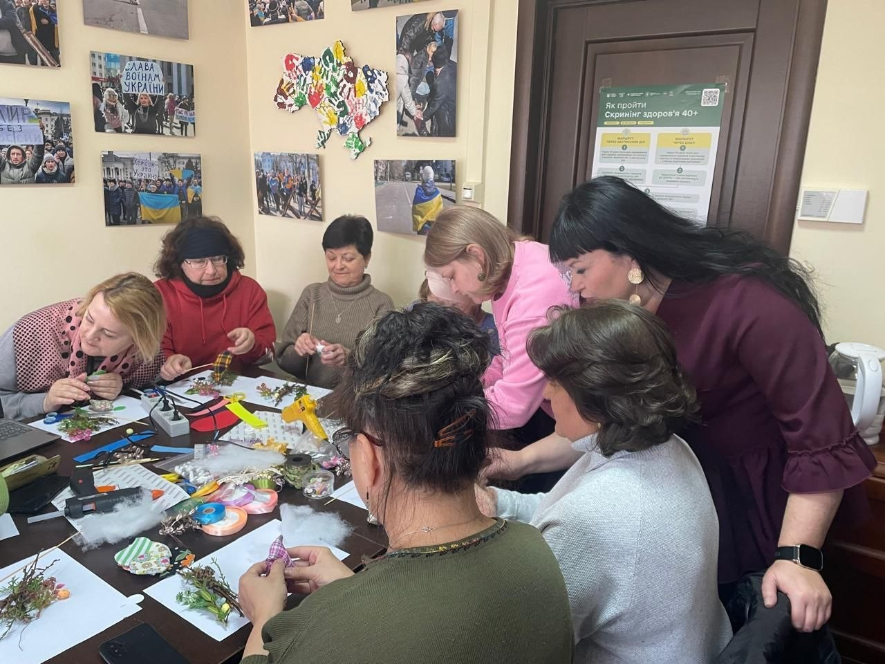 Група жінок за столом займається рукоділлям у світлій кімнаті. На столі лежать стрічки, клейові пістолети та заготовки для виробів; на стінах розміщено фотографії та мапа України.
