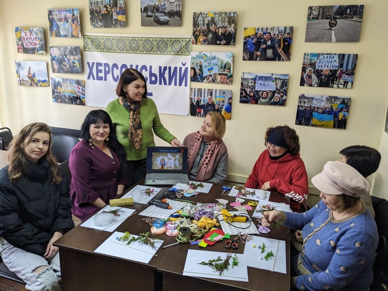 Група жінок за столом займається творчістю, виготовляючи вироби з природних матеріалів та стрічок. На стіні за ними — напис «Херсонський» та численні фотографії з патріотичною символікою.