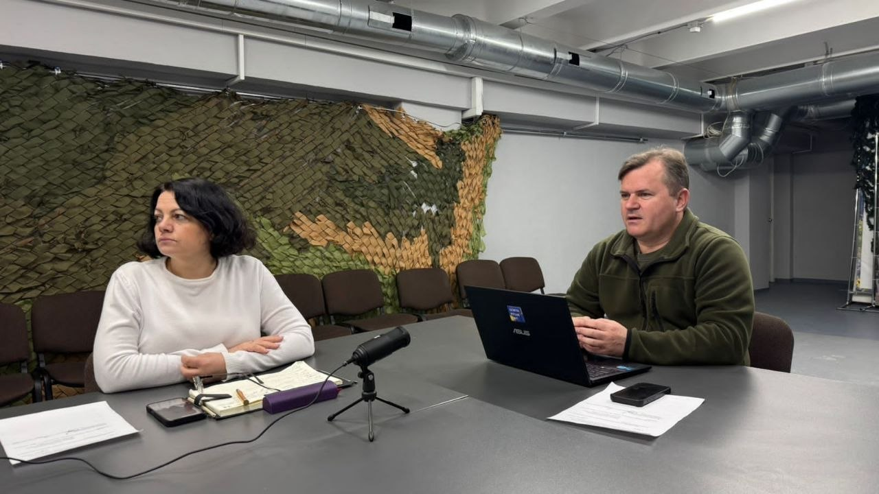 Чоловік та жінка сидять за столом під час робочої зустрічі або брифінгу. На столі перед ними ноутбук, мікрофон та нотатки; на фоні стіни — маскувальна сітка та елементи вентиляційної системи.