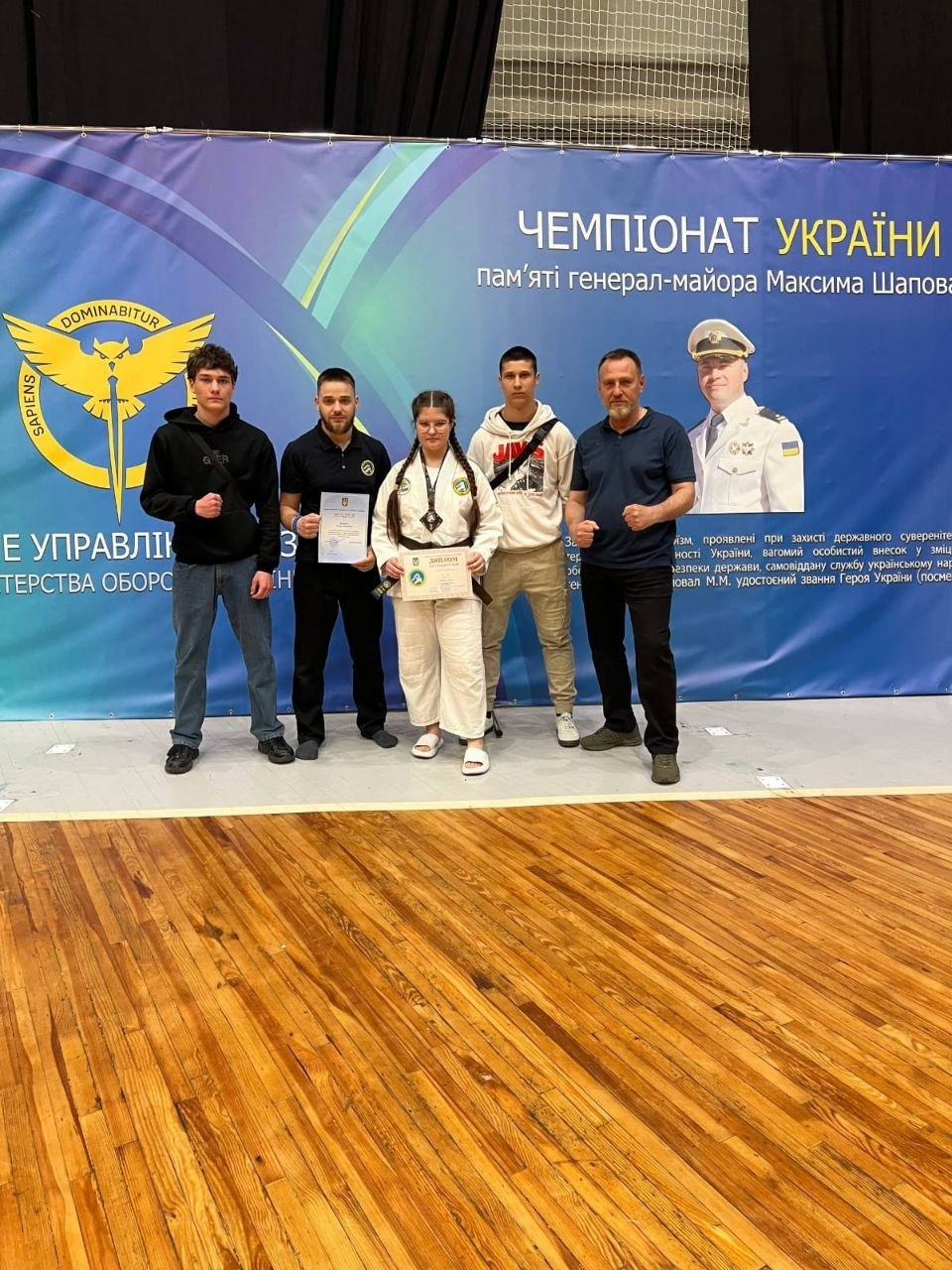 Група молодих людей та тренерів нагороджують спортсменку в кімоно на Чемпіонаті України пам’яті Максима Шаповала. У руках дівчини медаль та грамота; на фоні — великий банер із портретом героя та символікою Головного управління розвідки.