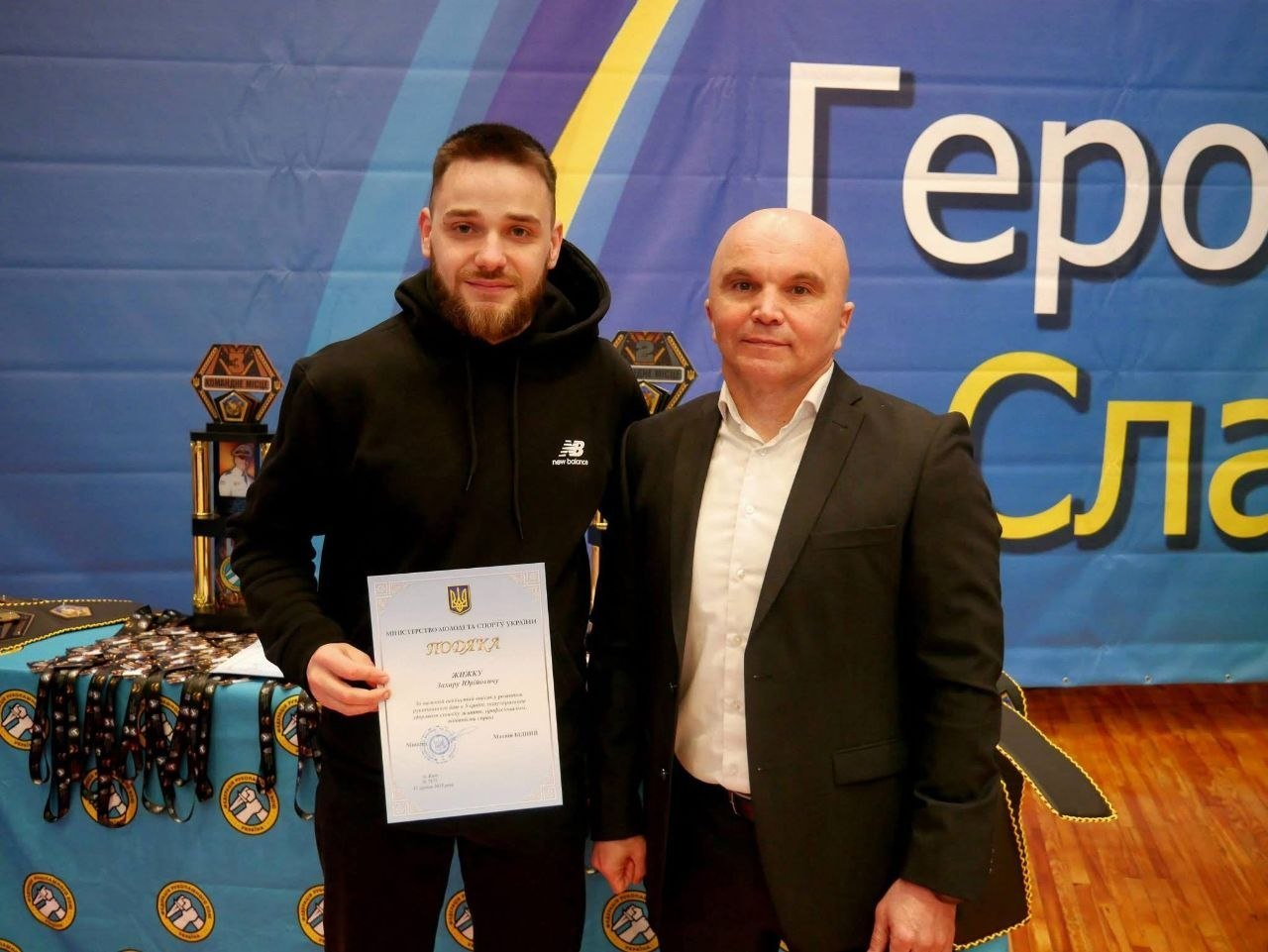 Двоє чоловіків позують на фоні синього банера з патріотичним написом. Чоловік зліва тримає в руках офіційну грамоту-подяку від Міністерства молоді та спорту України. На столі поруч розкладено численні медалі та кубки спортивного заходу.