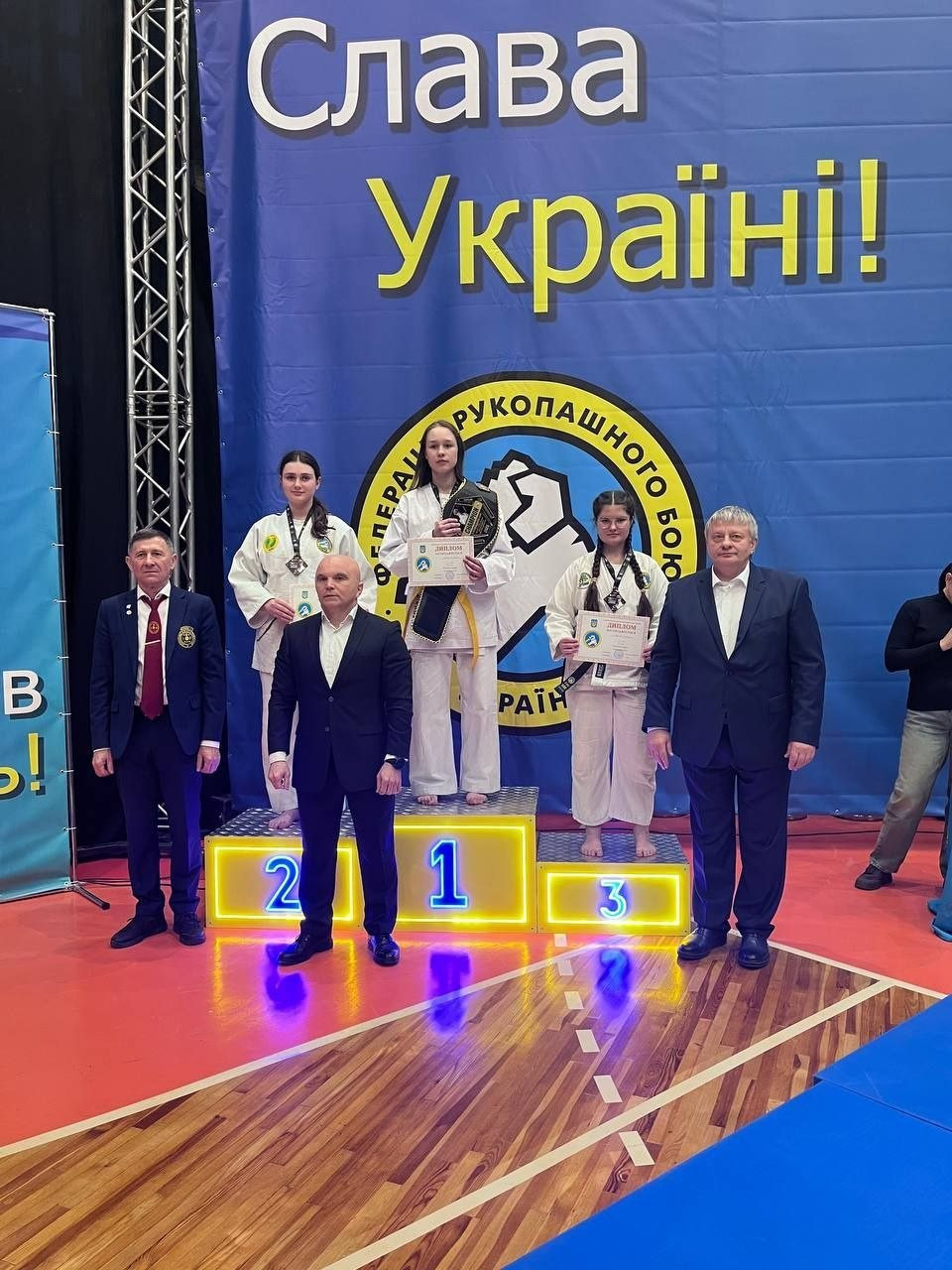 Троє дівчат-спортсменок у кімоно з медалями та грамотами стоять на призовому подіумі в спортивній залі. Поруч із ними троє чоловіків у костюмах; на фоні — синій банер із написом «Слава Україні!» та емблема федерації рукопашного бою.