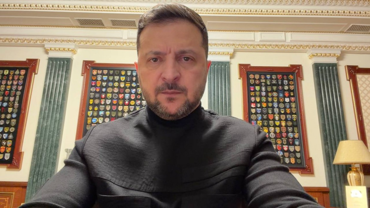 Чоловік у чорному светрі звертається до глядачів на фоні кабінету з класичним інтер'єром. На стінах за його спиною розміщені стенди з численними військовими шевронами та емблемами підрозділів.