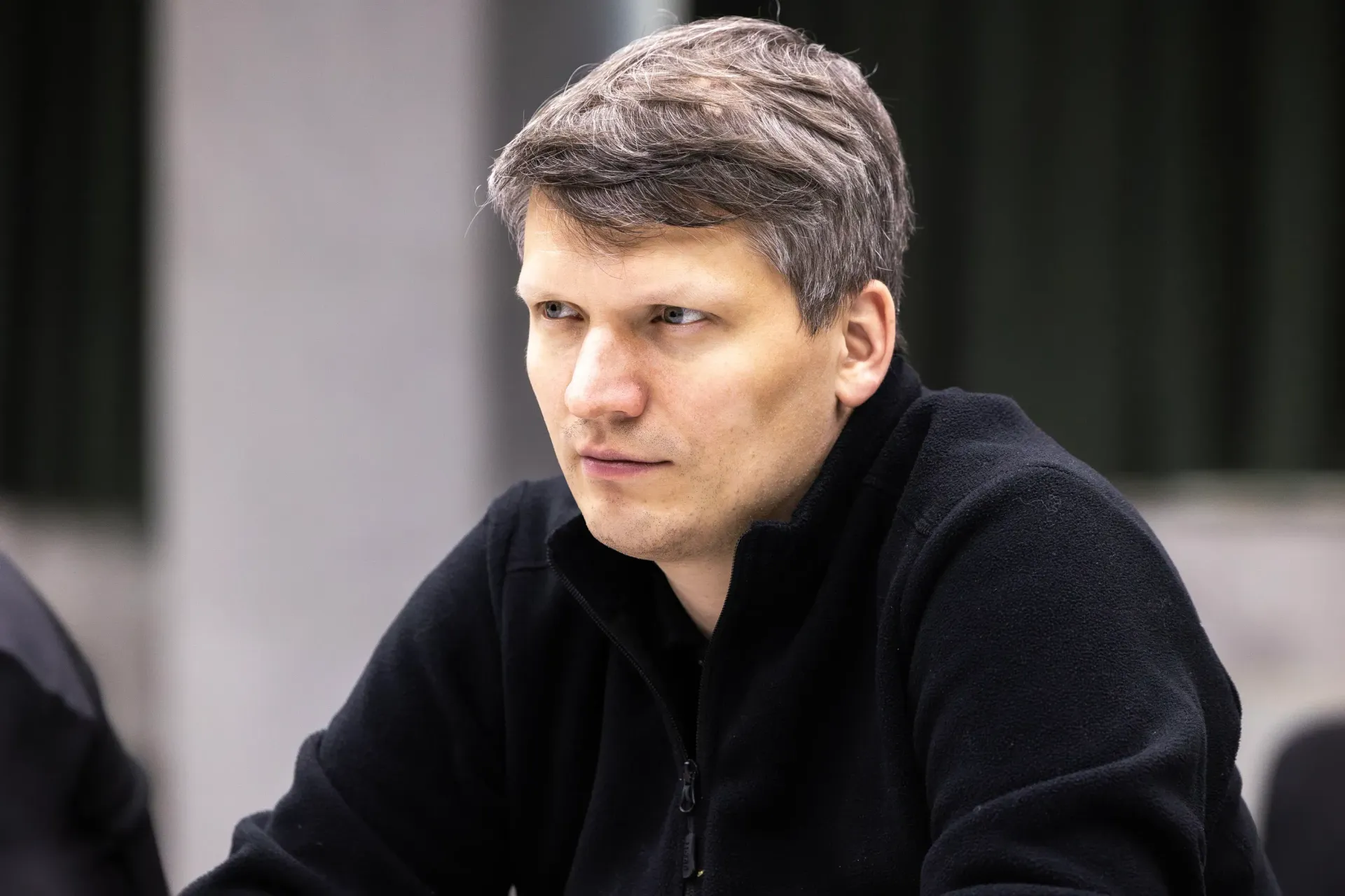 Портрет чоловіка середнього віку з коротким русявим волоссям, який дивиться вбік із серйозним або зосередженим виразом обличчя. Він одягнений у чорну флісову кофту на блискавці. Фон розмитий, що підкреслює постать чоловіка.