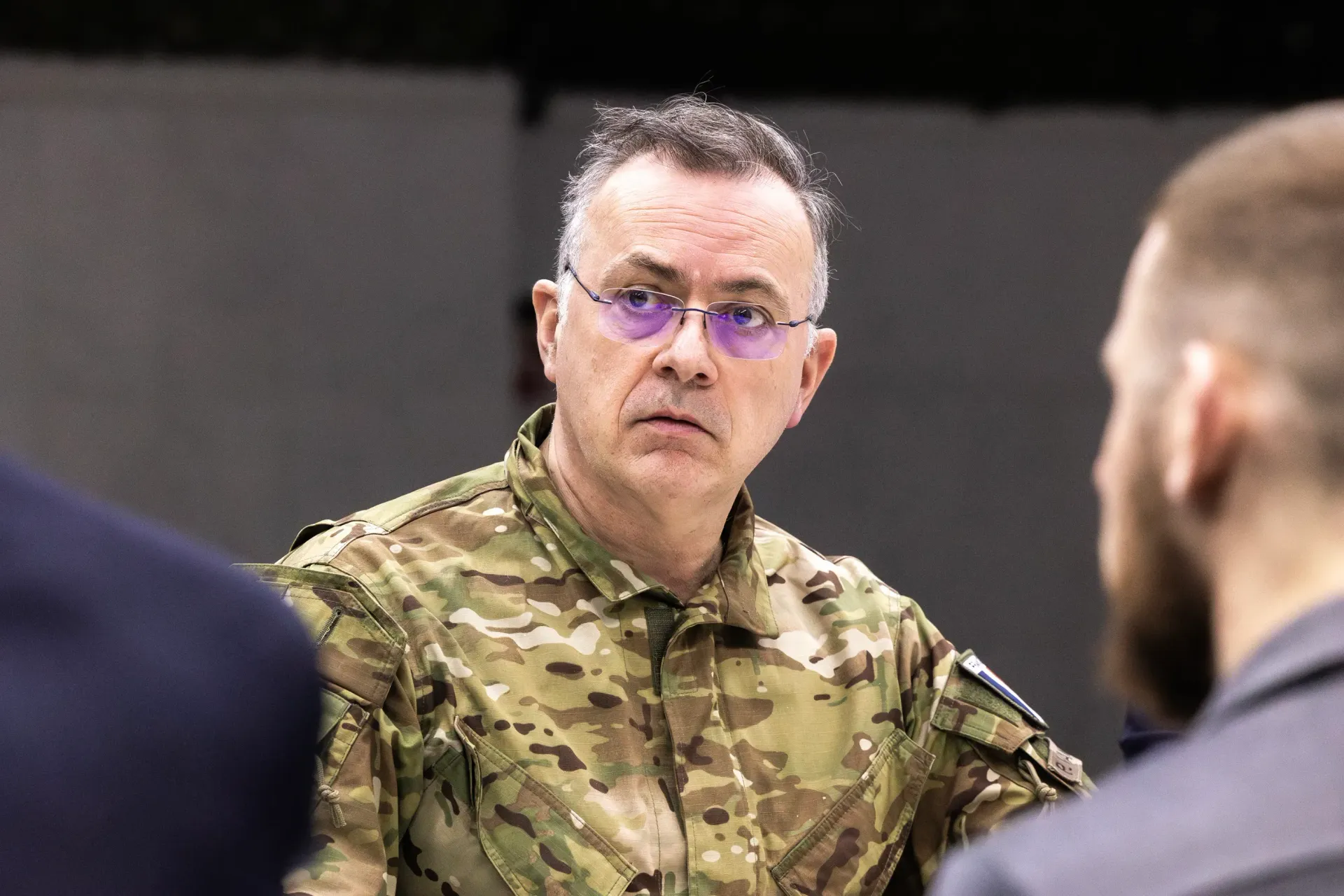 На фото зображений чоловік середнього віку в окулярах та військовій формі з камуфляжним візерунком. Він дивиться вбік із зосередженим виразом обличчя, перебуваючи в приміщенні серед інших людей, чиї постаті видно на передньому плані розмитими.