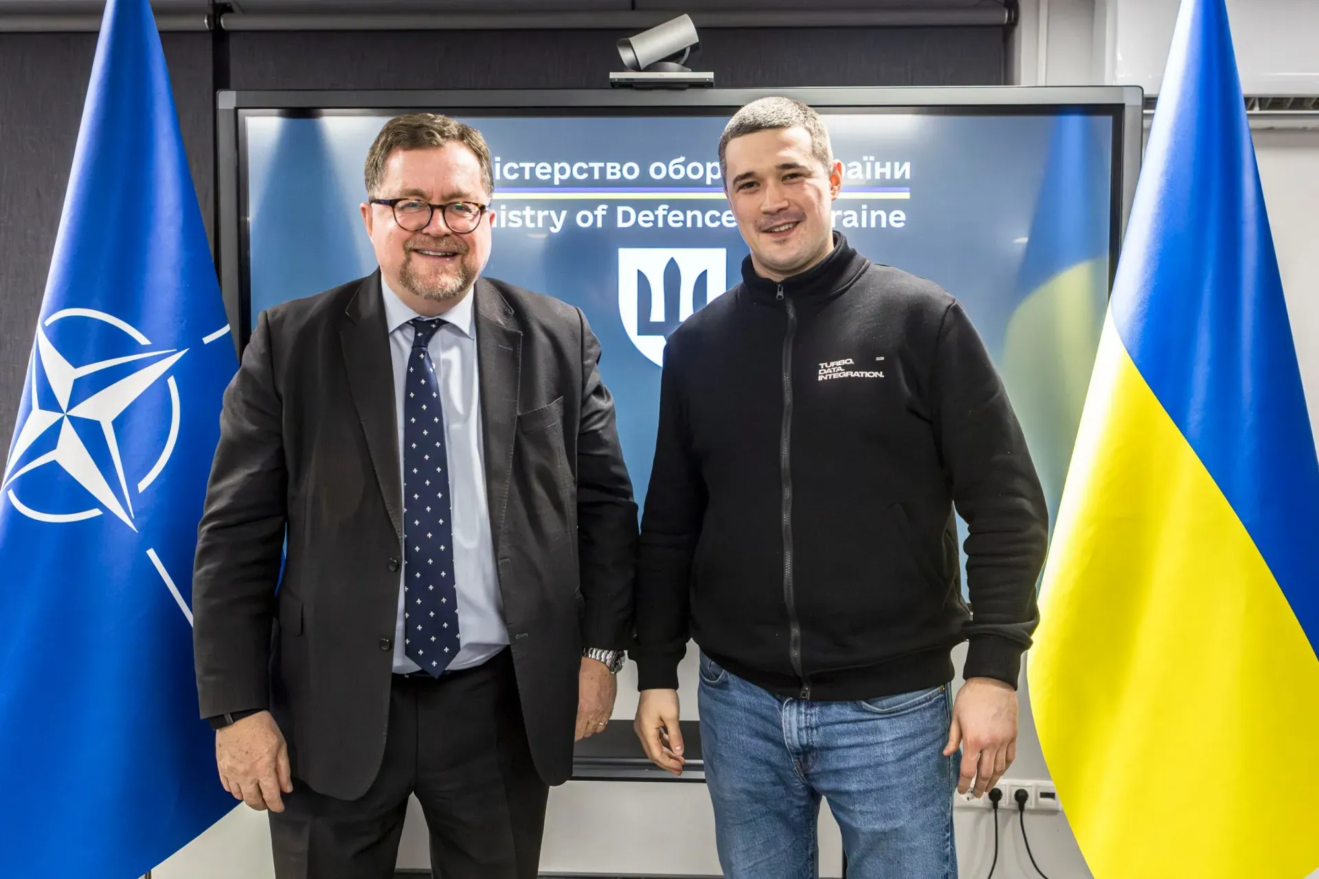 Двоє чоловіків у діловому та повсякденному одязі стоять між прапорами НАТО та України на фоні екрана з емблемою Міністерства оборони.