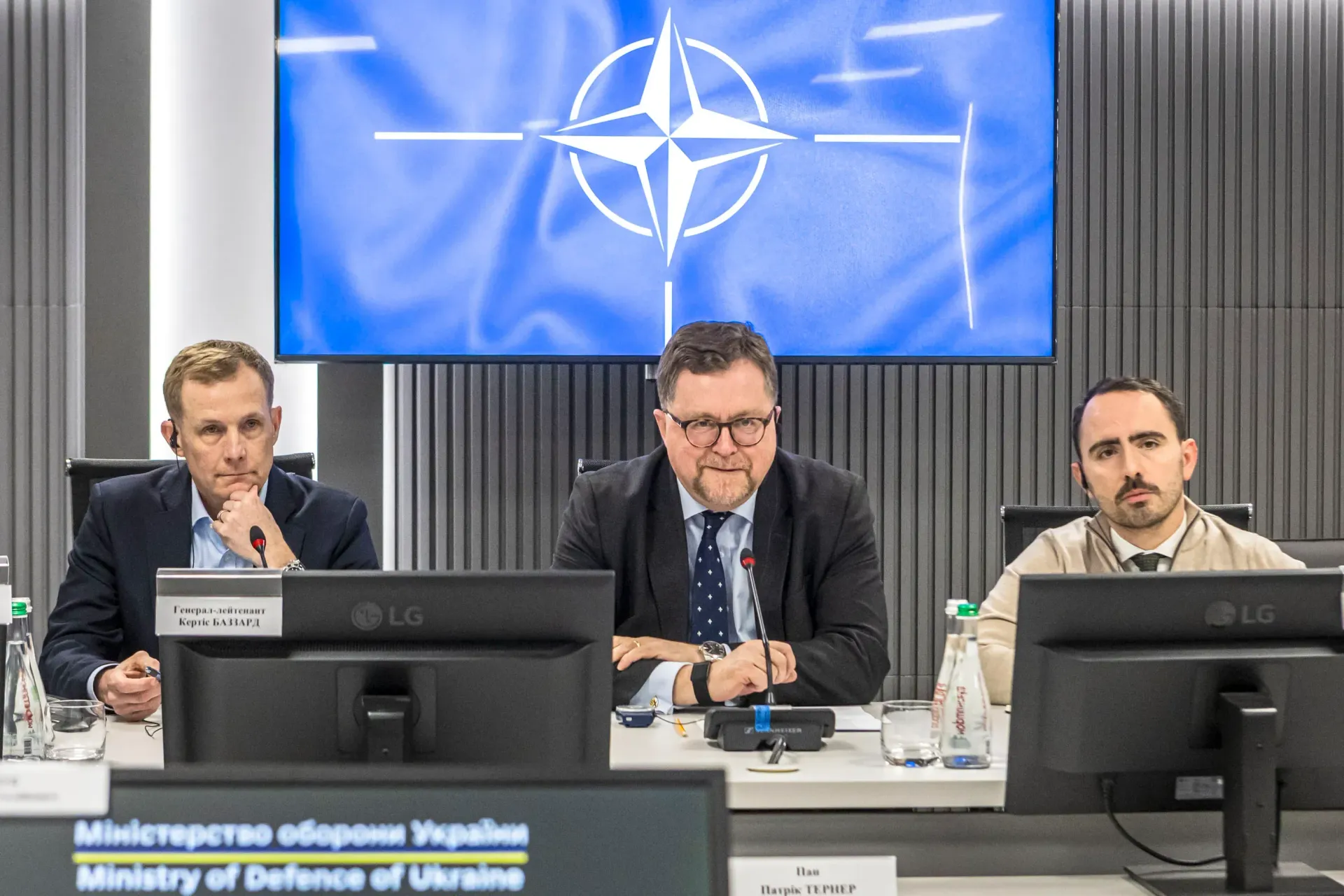 Троє чоловіків у діловому одязі сидять за столом під час офіційної зустрічі на фоні великого екрана з емблемою НАТО. Перед ними встановлені монітори та мікрофони, а на передньому плані видно табличку з написом «Міністерство оборони України».