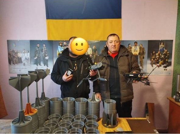 На фото двоє чоловіків у приміщенні з українським прапором демонструють дрони та компоненти до них. Обличчя одного з них приховано графічним символом. На передньому плані — численні деталі, виготовлені методом 3D-друку.