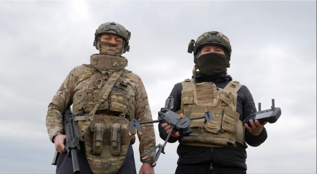 Двоє військових у захисному спорядженні та балаклавах стоять просто неба. Один тримає автоматичну зброю, інший — квадрокоптер та пульт керування ним.