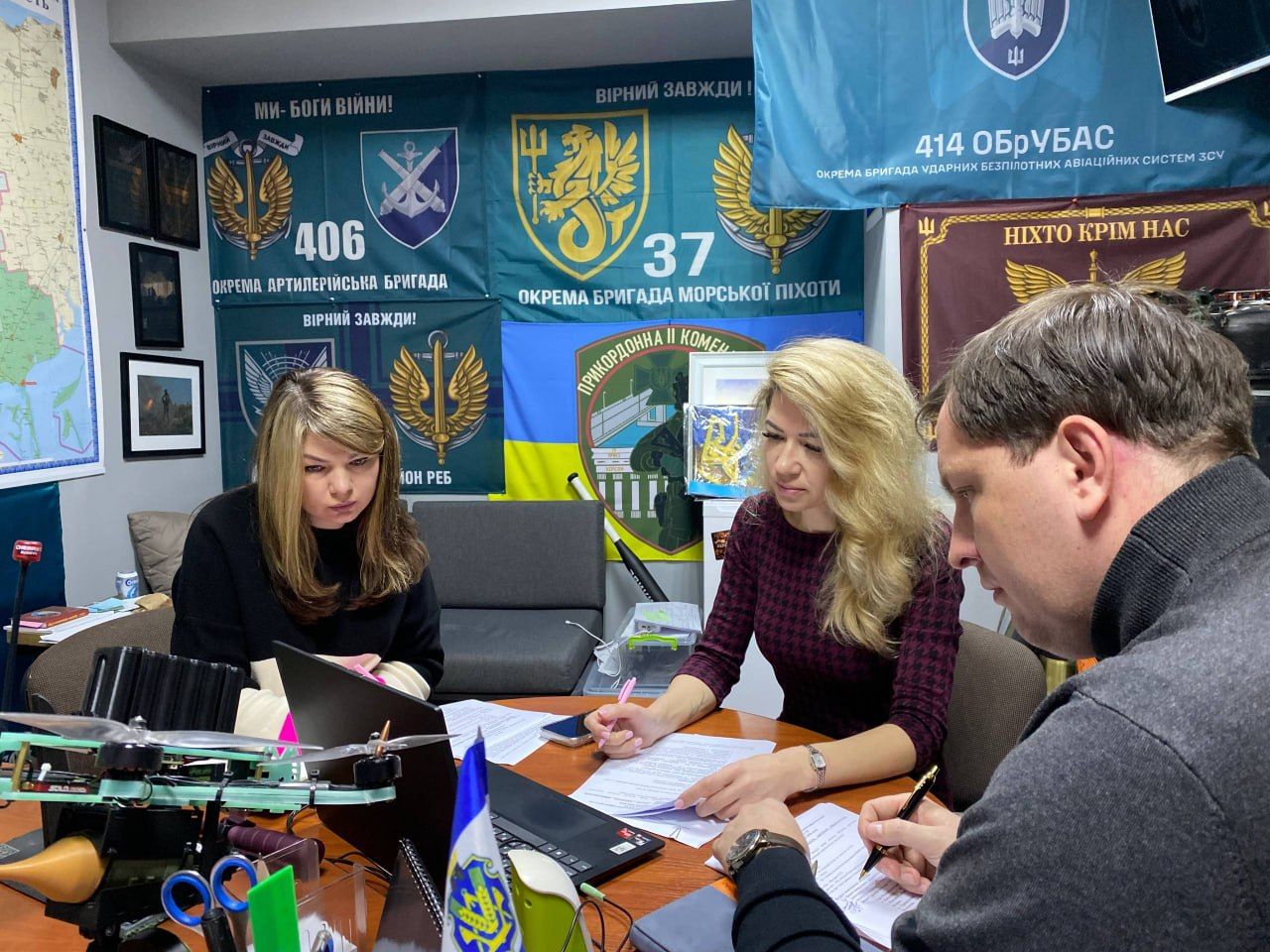 На фото троє людей у приміщенні з українською військовою символікою на стінах зосереджено працюють із документами за столом, на якому стоять ноутбук та канцелярське приладдя.