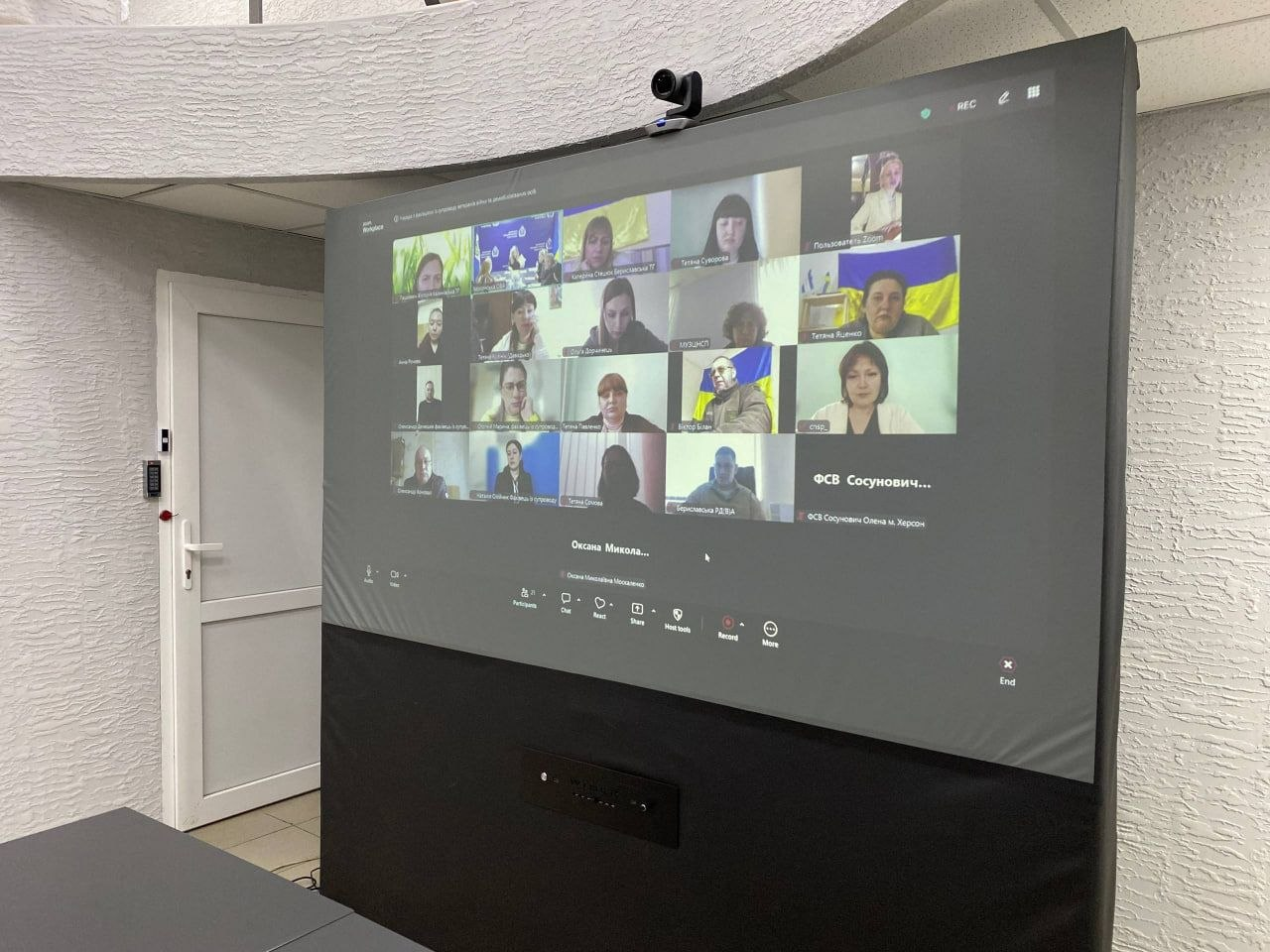 Великий екран у приміщенні, на якому транслюється онлайн-нарада з багатьма учасниками у форматі відеоконференції.