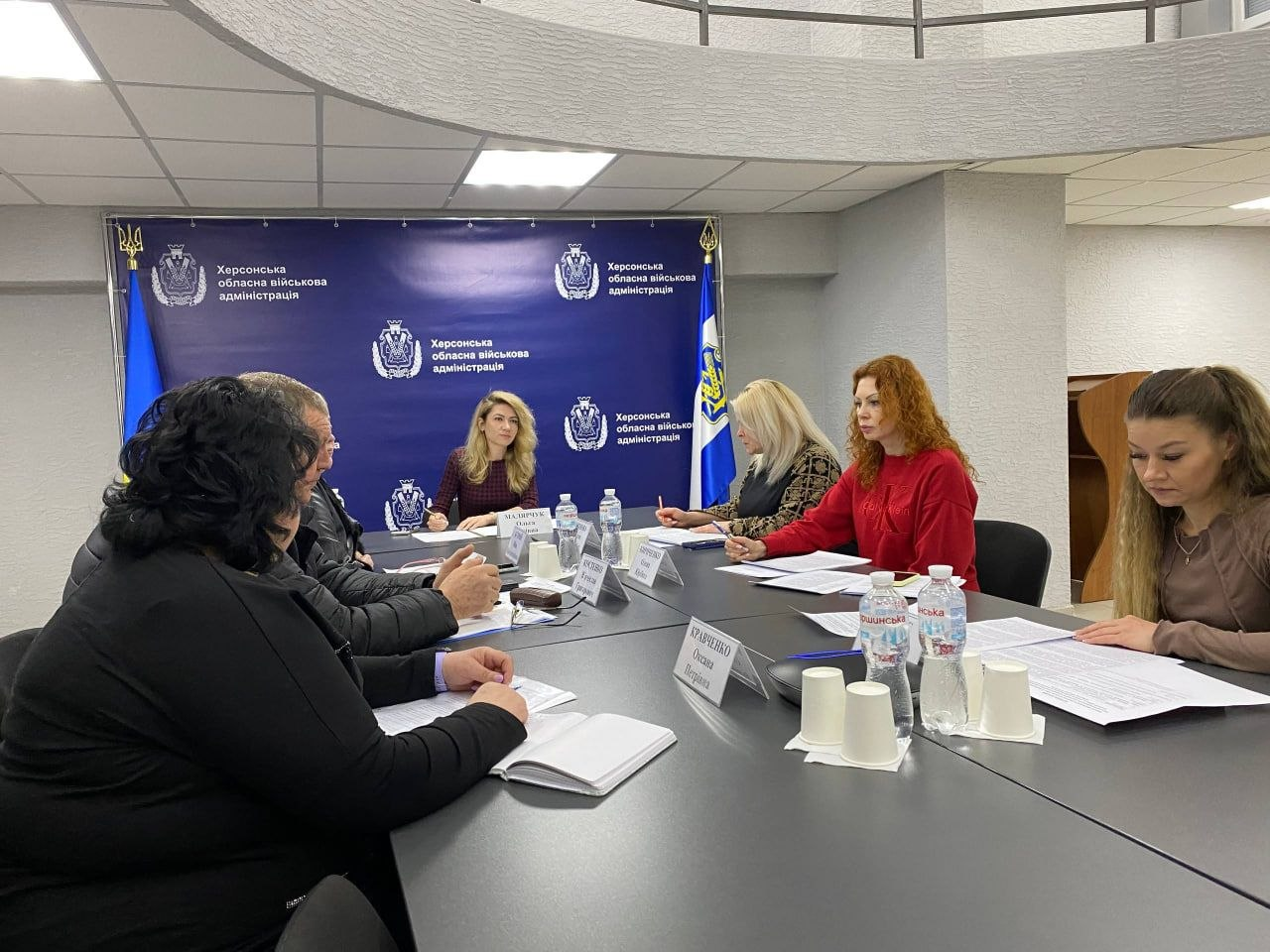 Учасники офіційної наради за робочим столом із документами на фоні банера з державною символікою.