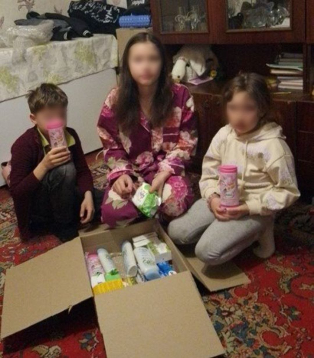 Троє дітей сидять на підлозі біля відкритої коробки з наборами засобів гігієни та побутової хімії.